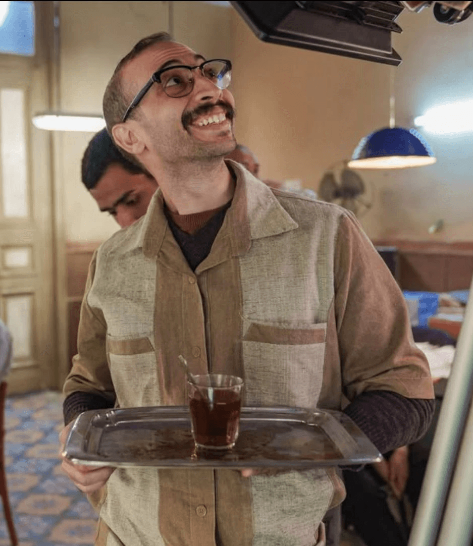 A smiling man with glasses and a mustache holds a tray with a glass of tea. He's wearing a collared shirt and is looking upward.