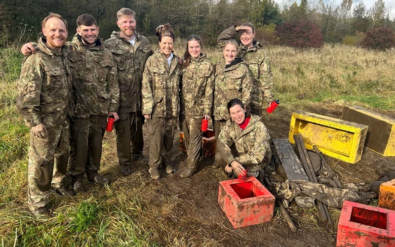 A group wearing camouflage clothing stands outdoors in a muddy field during a practical activity. Equipment boxes and tools nearby suggest a hands‑on training or teamwork exercise in a rural setting.