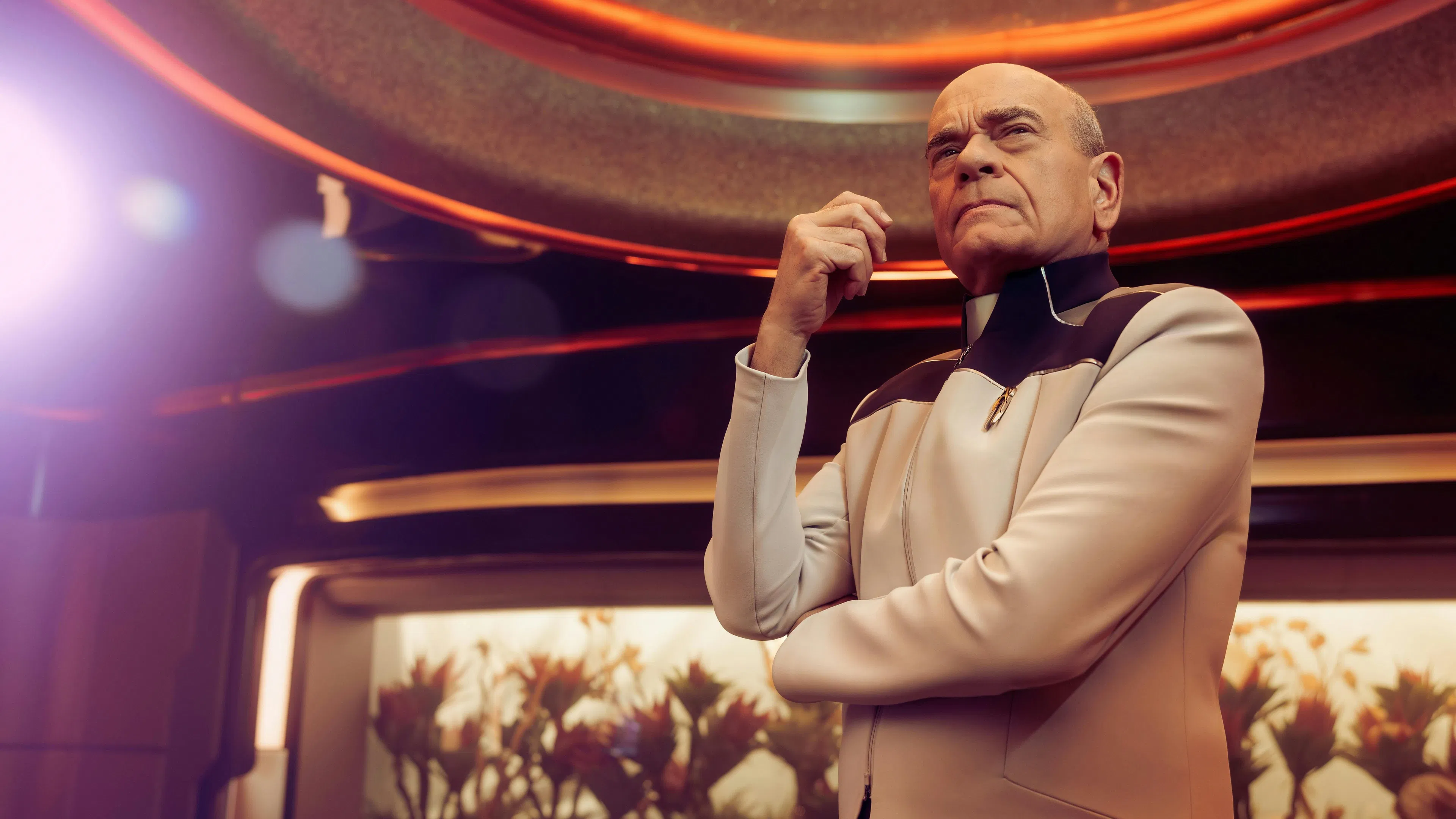 A senior man in futuristic attire stands thoughtfully under warm, ambient lighting, surrounded by sleek, modern décor and illuminated plants, suggesting a sci-fi setting.