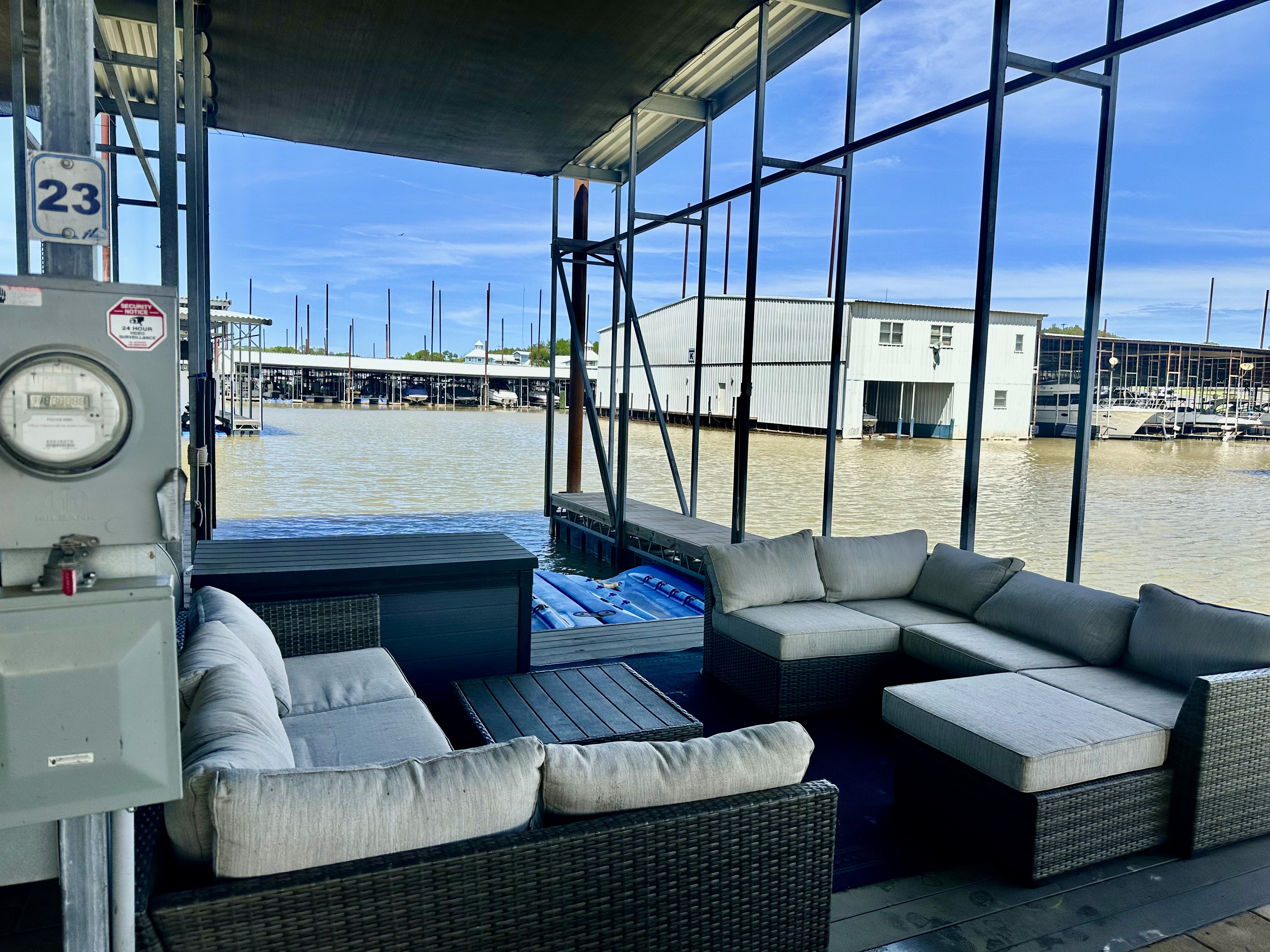Modern outdoor seating area with gray cushioned wicker furniture on a dock overlooking a marina with multiple boat storage buildings; the dock is sheltered by a metal roof and framed structure, enhancing the waterfront ambiance.