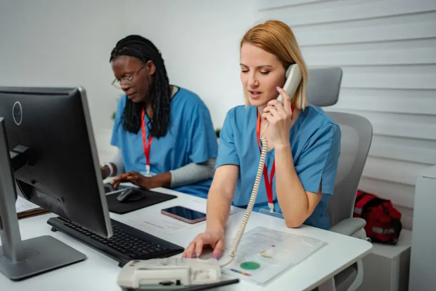 AI assistant for healthcare shown as two medical workers in blue scrubs managing phone calls and computer tasks at a desk.