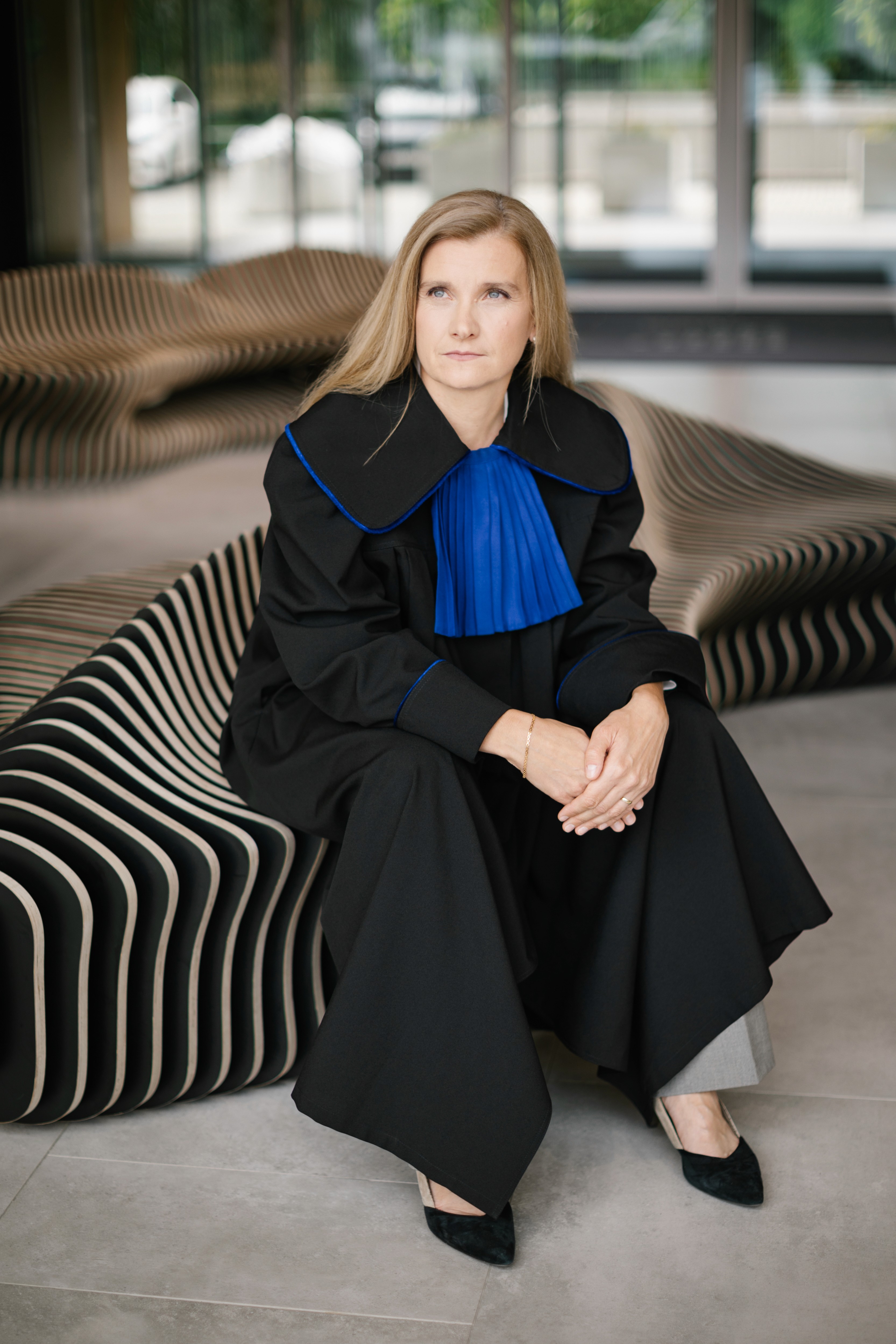 obieta o blond włosach, ubrana w czarną szatę z niebieskim plisowanym kołnierzem, siedzi na nowoczesnej, pasiastą ławce w pomieszczeniu, patrząc lekko w bok z zamyślonym wyrazem twarzy.