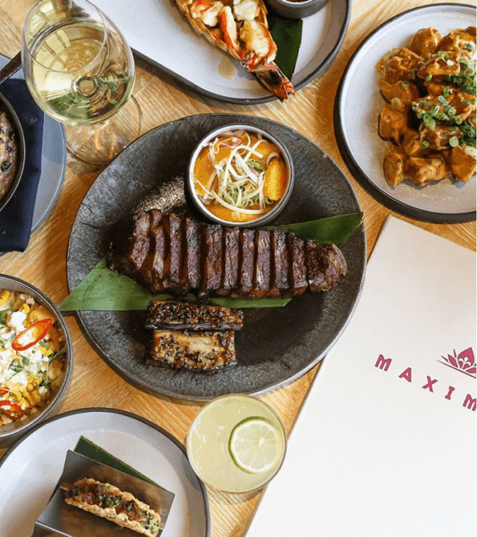 Plated steak with sides, tacos, seafood, cocktails, and a menu labeled Maximon arranged on a wooden table.
