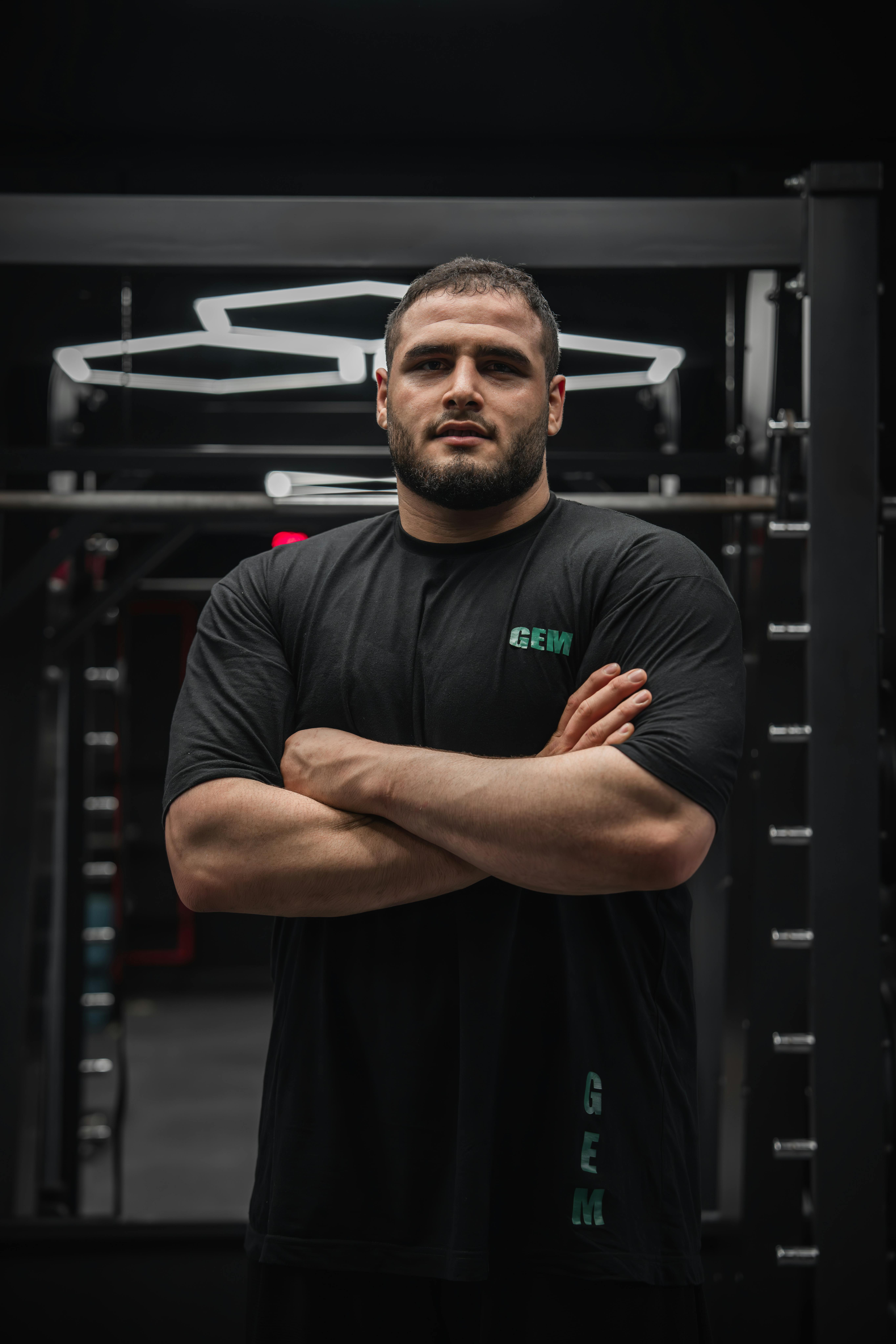 A man with a muscular build and beard stands confidently with arms crossed, wearing a black GEM-branded T-shirt, in a dimly lit modern gym with metal equipment in the background.