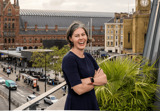 A portrait of New Practice Lab's Executive Director, Tara McGuinness, standing and smiling on a balcony overlooking a busy street and ubran background.