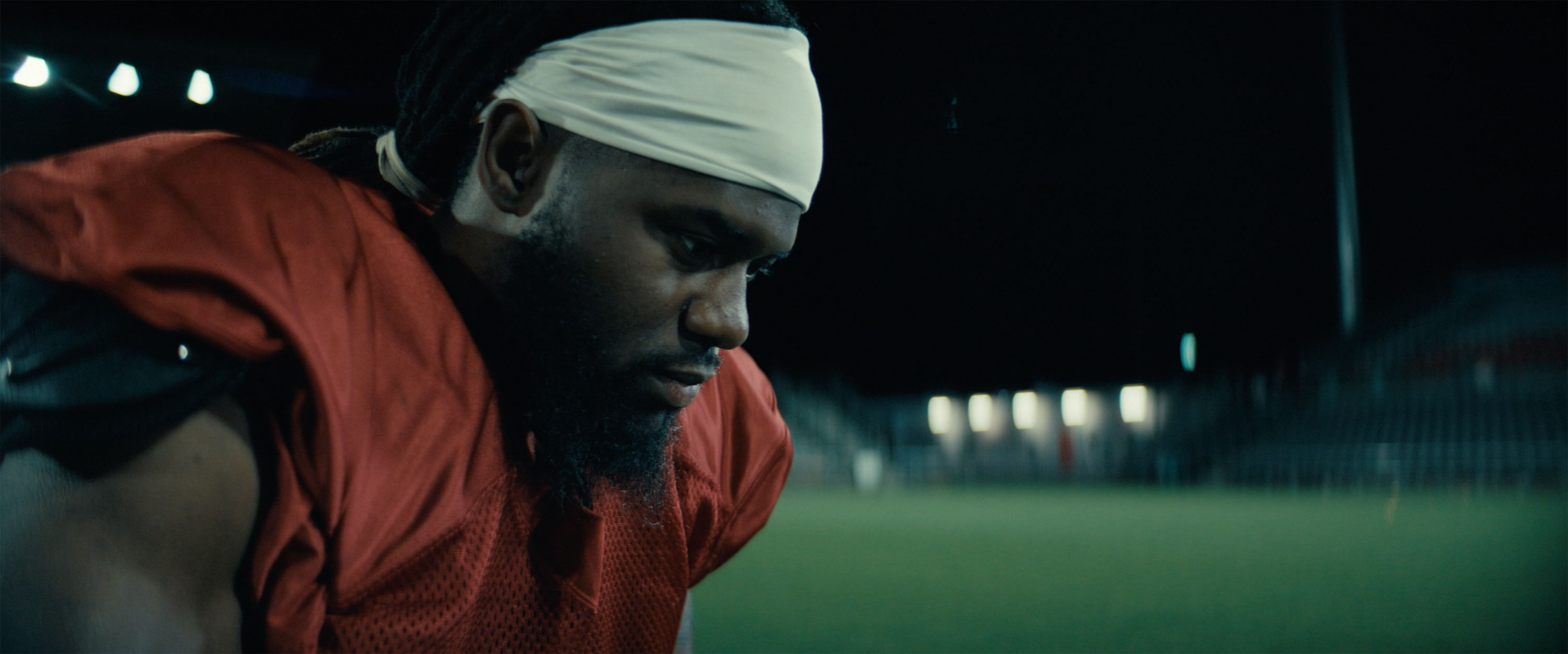 Close-up of a football player in a red jersey and headband, looking down on a dimly lit stadium field.