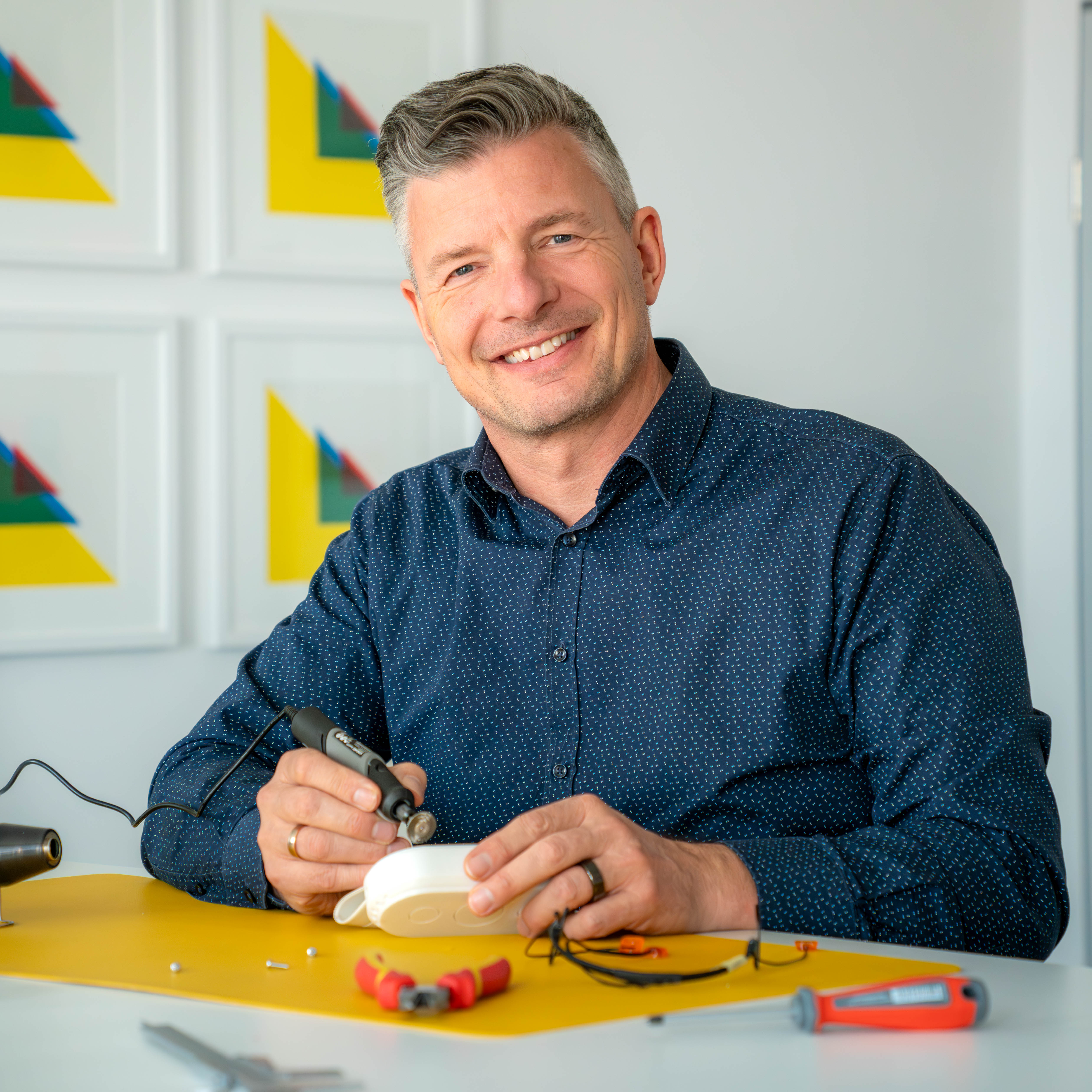 Mann mit blauem Hemd sitzt an einem Tisch und hält ein elektrisches Werkzeug über einem weißen Objekt