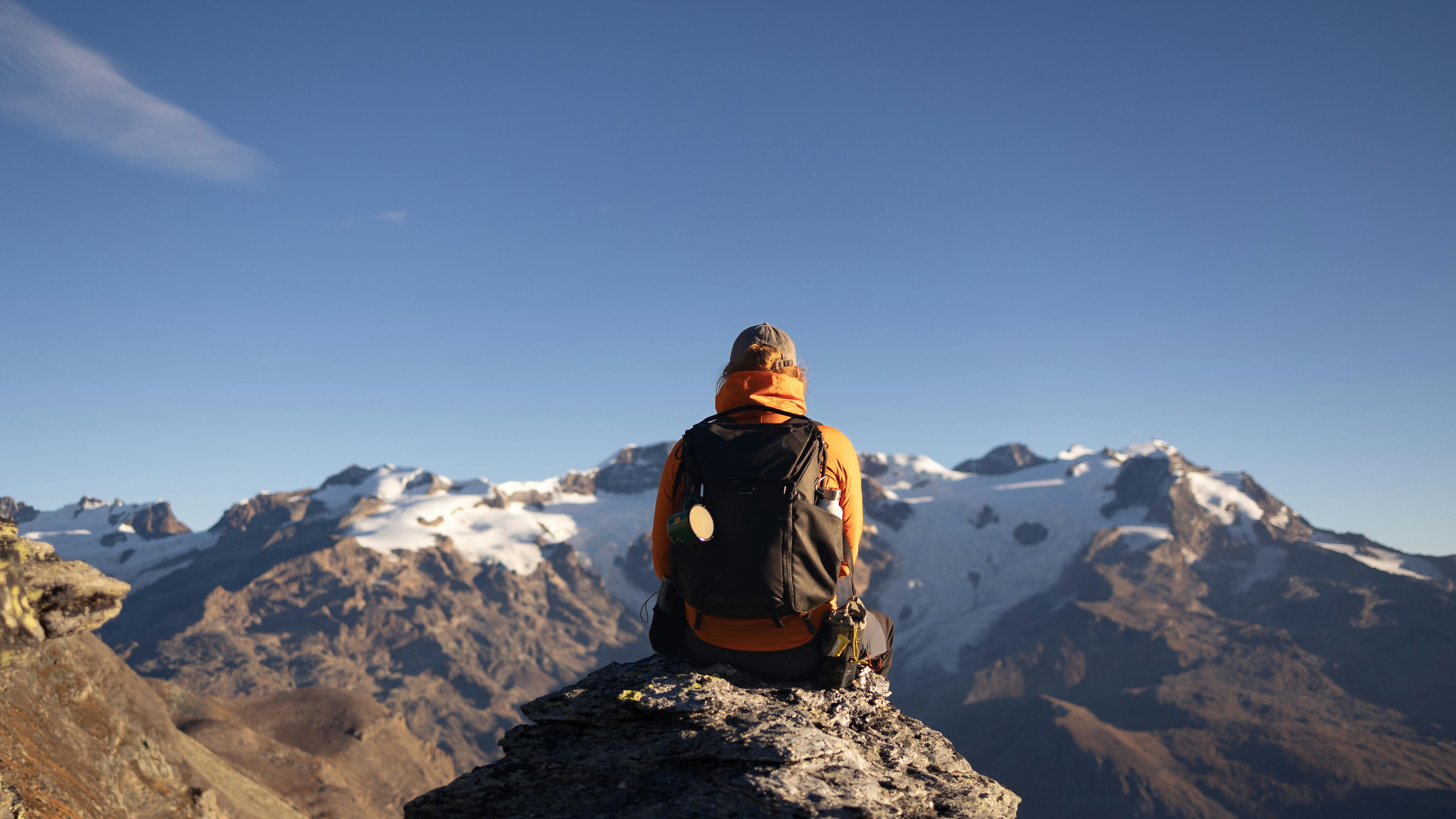 A man sits peacefully on a rocky mountain peak, gazing out at the expansive view of rolling hills and valleys bathed in soft, natural light.