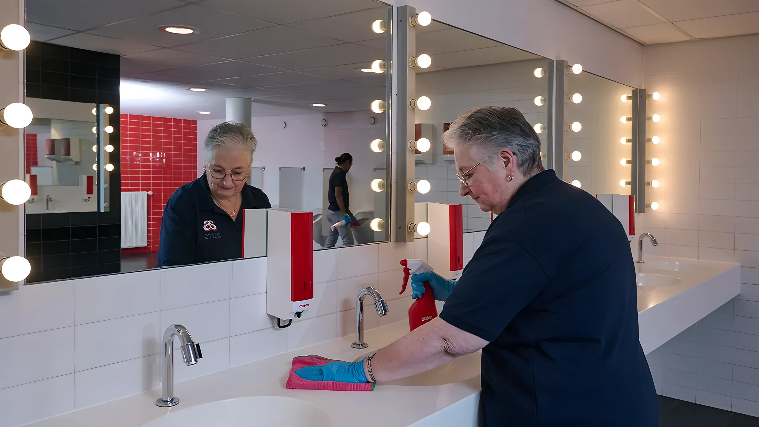 Twee schoonmaakmedewerkers aan het werk bij de wastafels van een kleedkamer met spiegellampen
