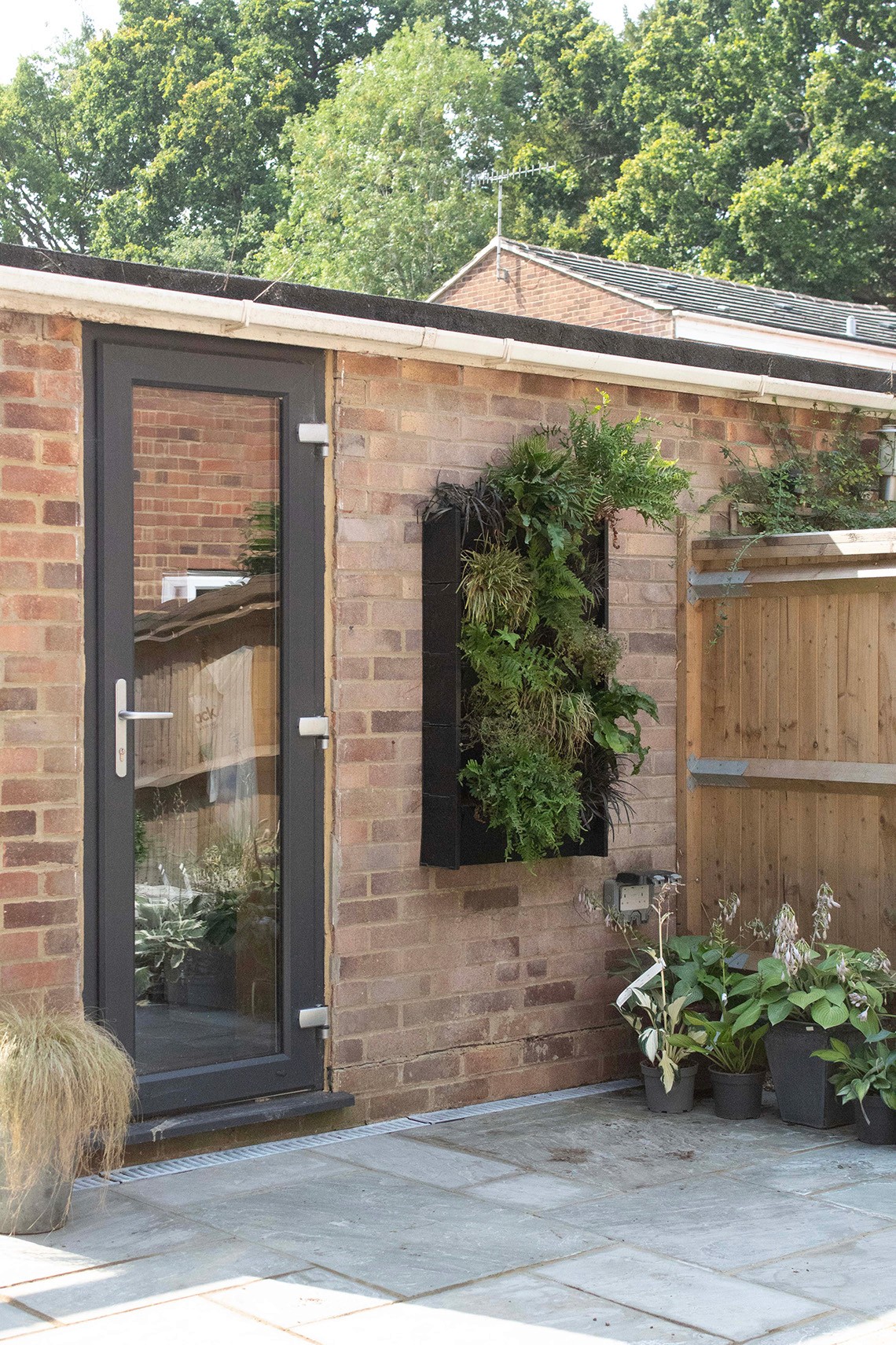 A small brick structure with a dark door and a window filled with greenery, surrounded by a path and fence.