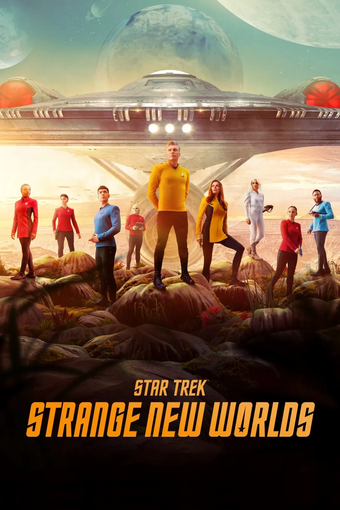 Image of a science fiction scene featuring a group of people in futuristic uniforms standing on a rocky alien landscape, with a large spaceship hovering in the sky under a colourful, otherworldly sky; the scene is from "Star Trek: Strange New Worlds."