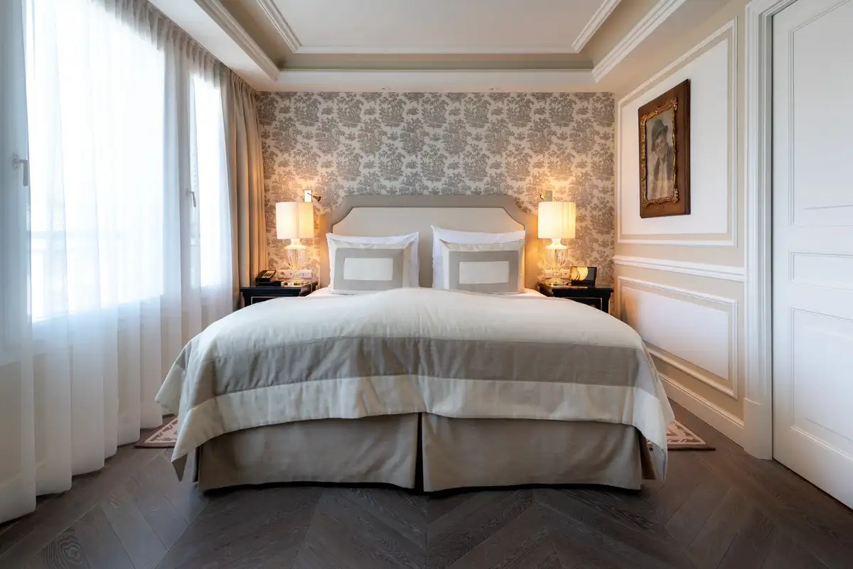 A large bed with white linens and decorative pillows sits against a patterned wallpaper wall in a bright hotel room.