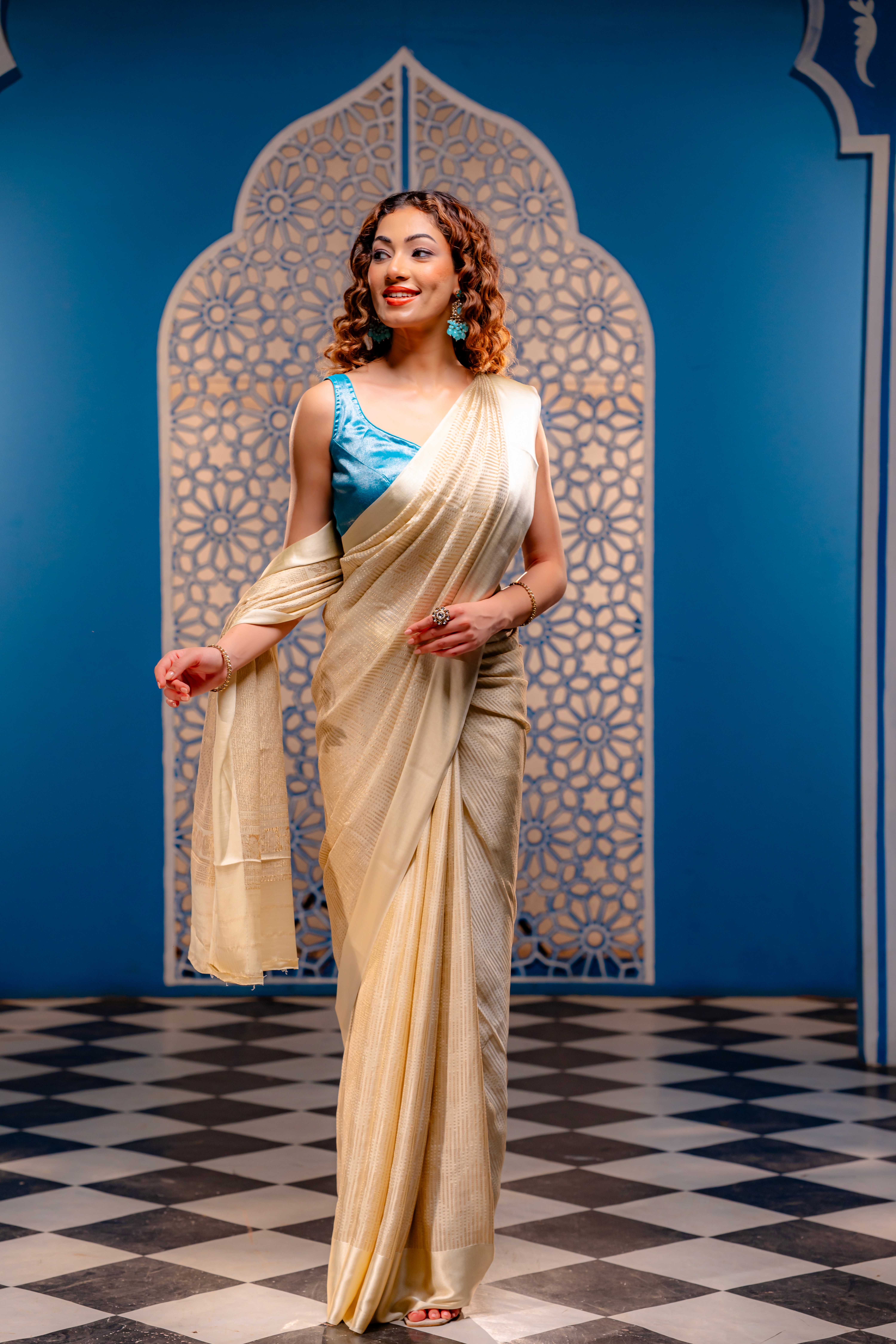 A woman wearing a cream saree paired with a blue blouse from That Saree Shop, standing before a striking blue wall adorned with beige Jaipur prints, creating a perfect blend of traditional elegance and artistic ambiance.