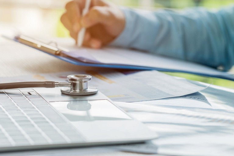 Healthcare professional filling out a checklist on a clipboard, with a stethoscope and laptop on the desk, representing ESG compliance for care homes
