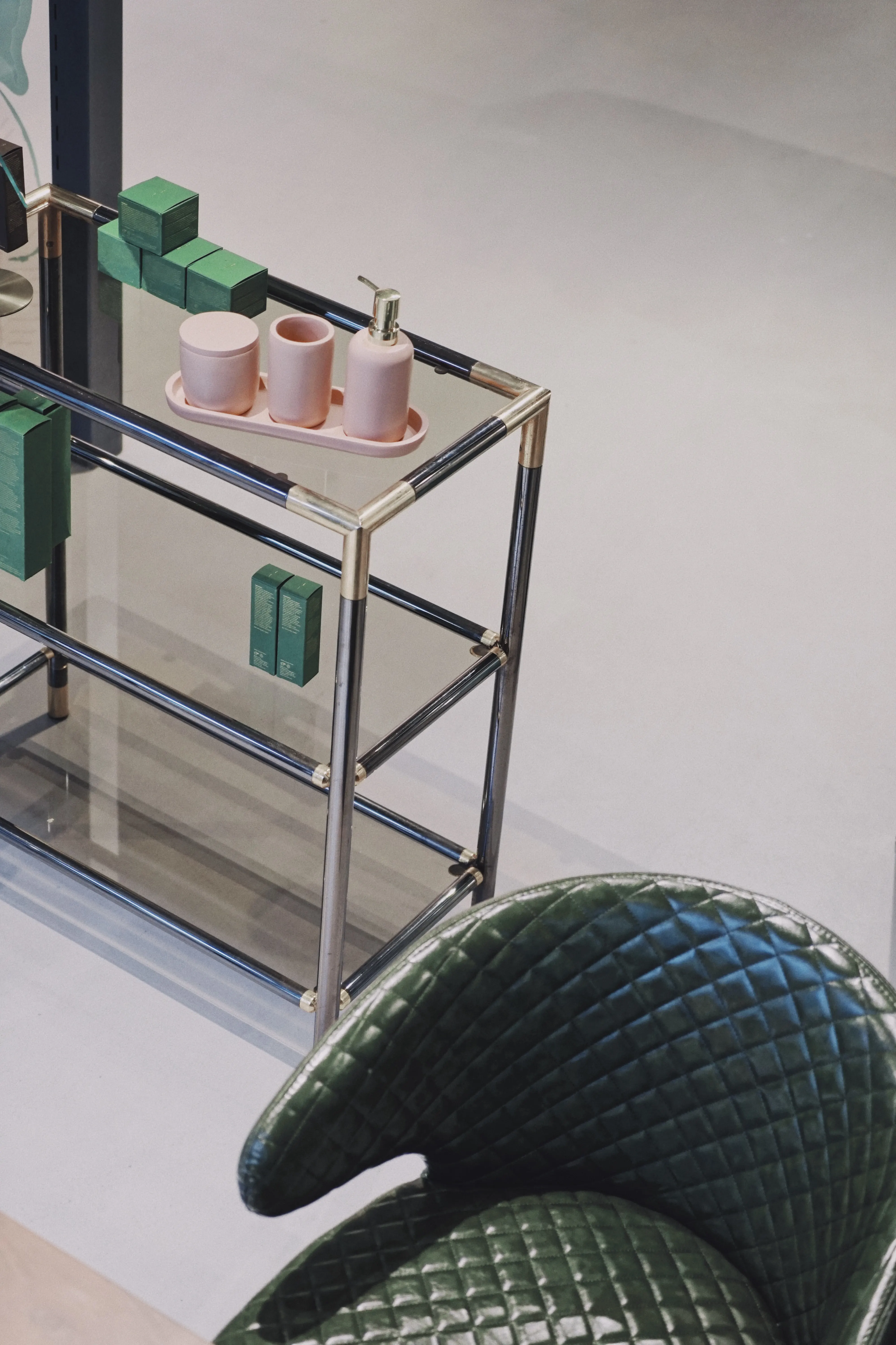 Detail at UZWEI FASHION HOTEL with a chrome-and-glass étagère holding pink cosmetics and green boxes, behind a quilted dark green leather chair.