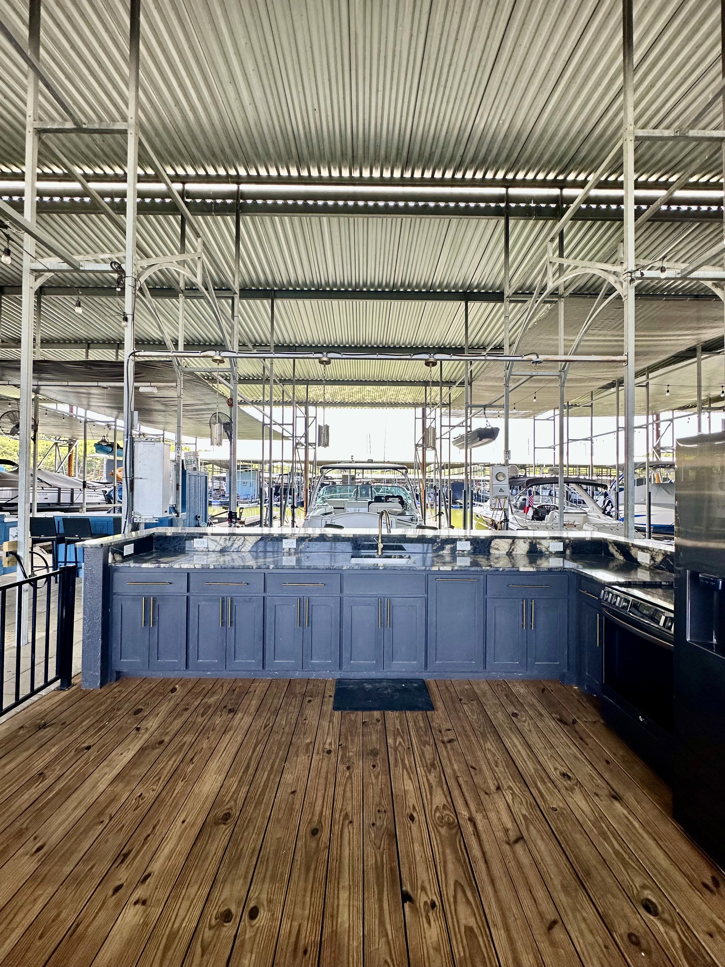 A modern outdoor kitchen on a wooden dock features a sleek blue cabinet with a marble countertop and a built-in stainless steel grill, set against a backdrop of a covered marina with boats and industrial metal beams.