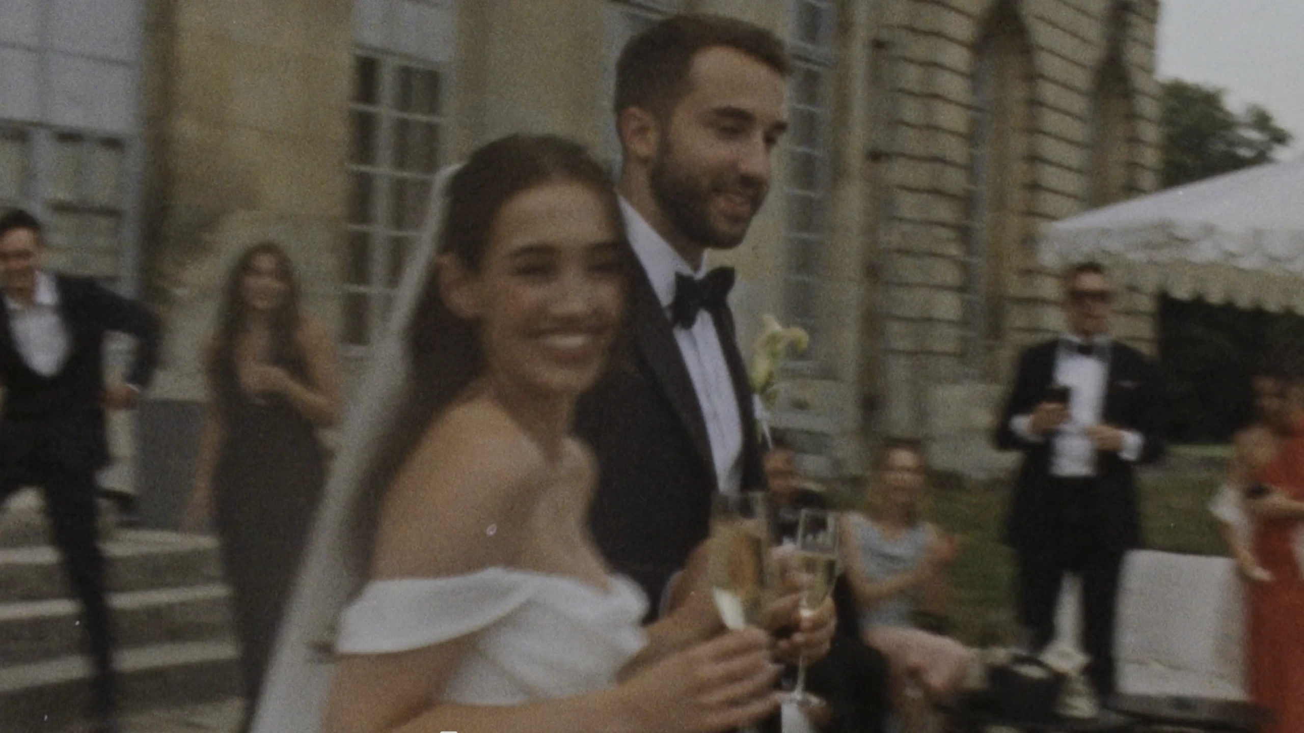 A joyful couple in wedding attire celebrates outside a historic stone building with guests in formal wear, holding glasses and surrounded by lush greenery.