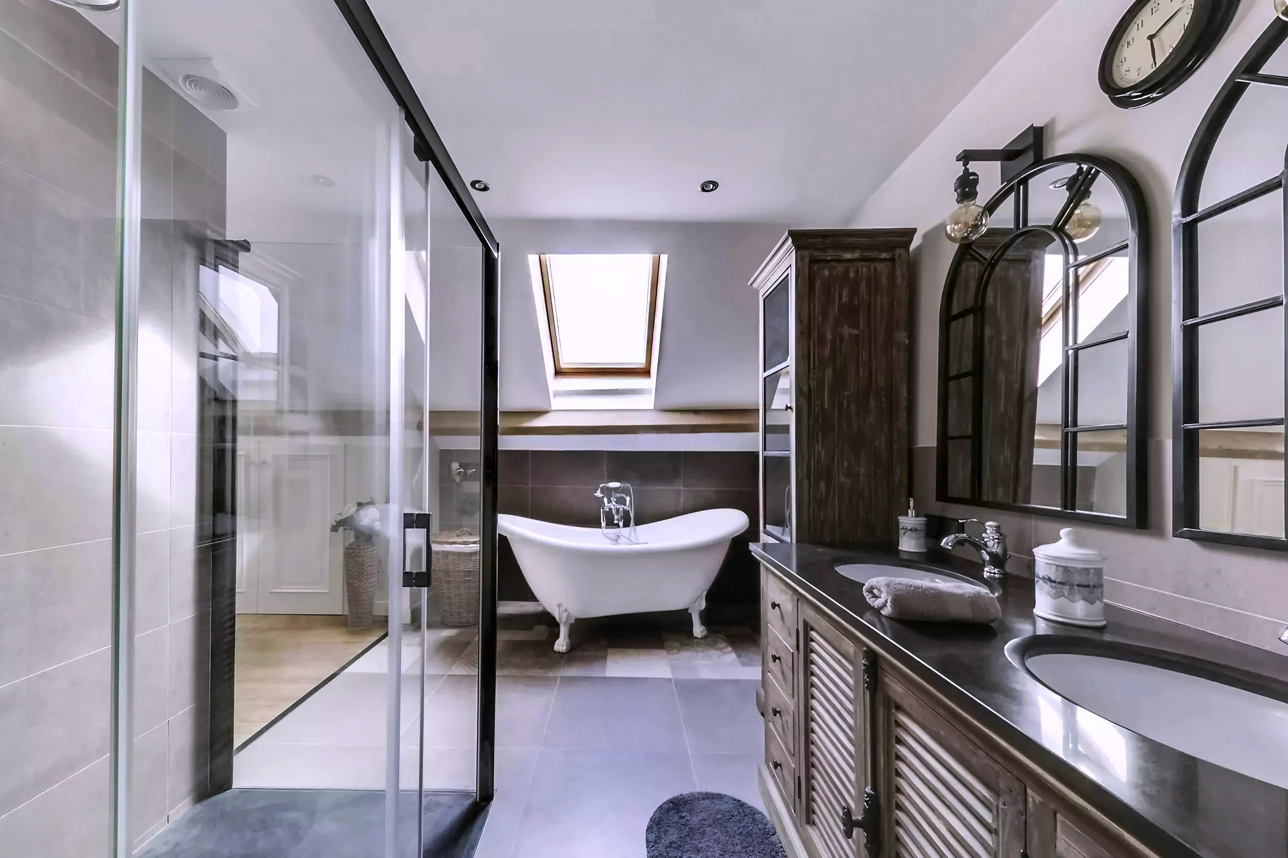 Transformation salle de bains Seine-et-Marne avec cabine douche verre, baignoire pieds blanche sous puits lumière et double lavabo bois foncé, rénovation complète.