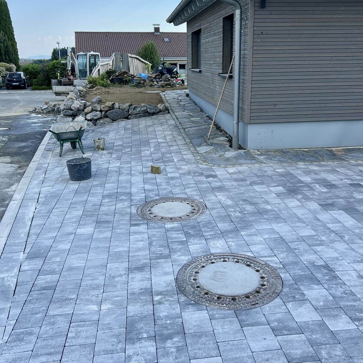 Baustelle zur Garten-Neuanlage mit verlegtem Pflasterweg, runden Schachtdeckeln und einem Schubkarren im Hintergrund, Gartengestaltung Klingenstein.