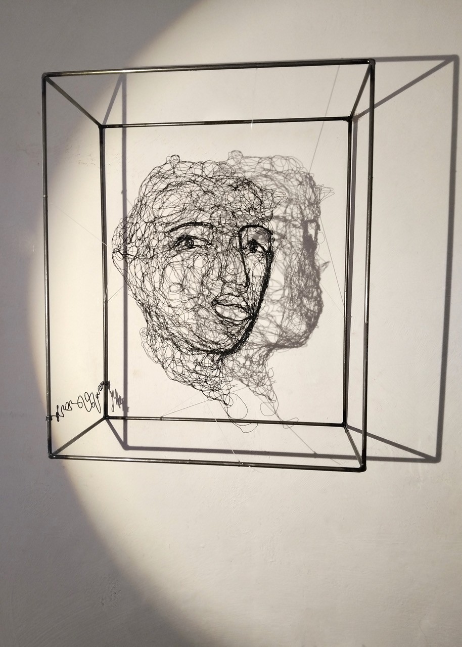Sculpture en fil de fer représentant le visage d'une femme dans un cadre en fil de fer, vue de loin avec son ombre projetée sur le mur