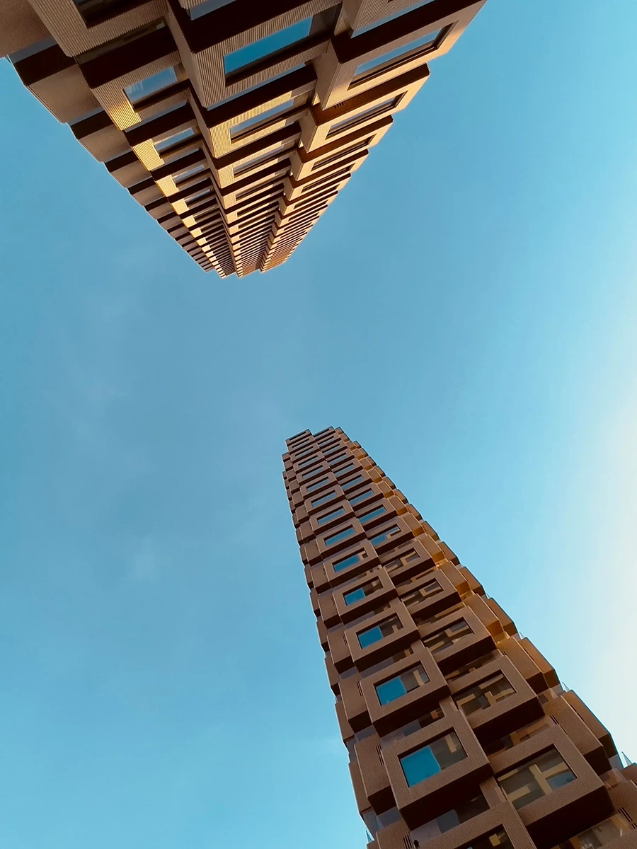 Aufwärtsperspektive von zwei hohen, modernen Gebäuden mit geometrischen Fassaden vor einem klaren blauen Himmel, die eine symmetrische und abstrakte architektonische Perspektive schaffen.