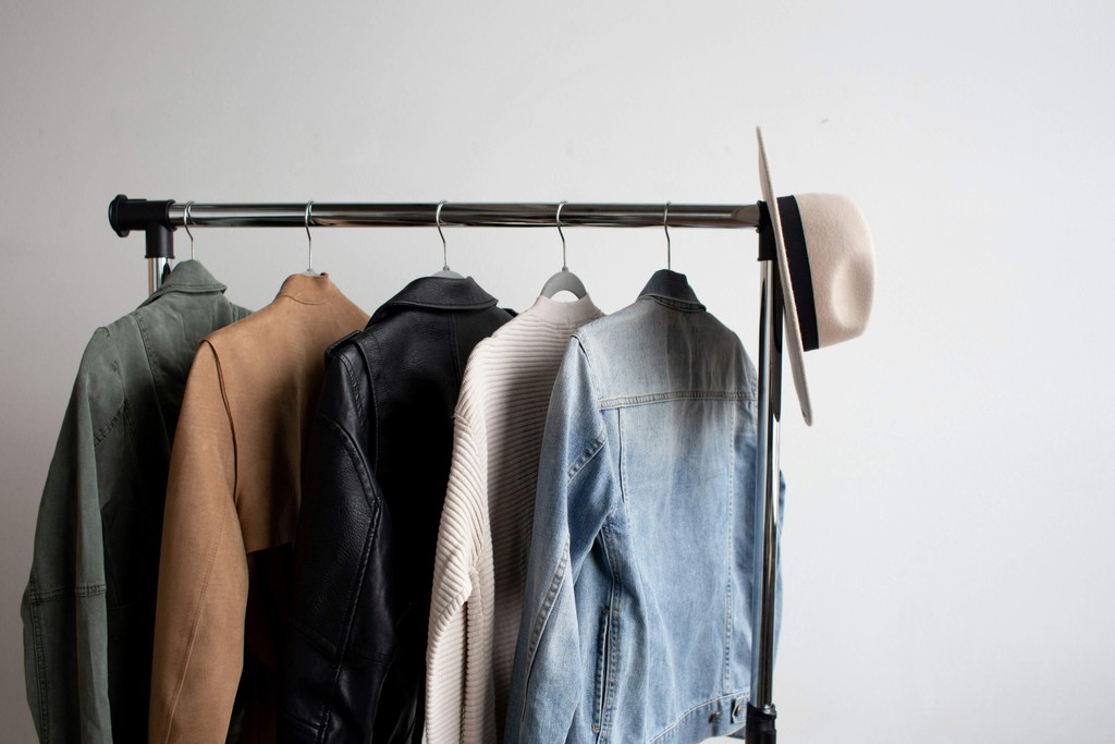 A clothing rack with several garments hanging on hangers and a wide-brimmed hat hanging on the end of the rack. The background is a plain white wall.