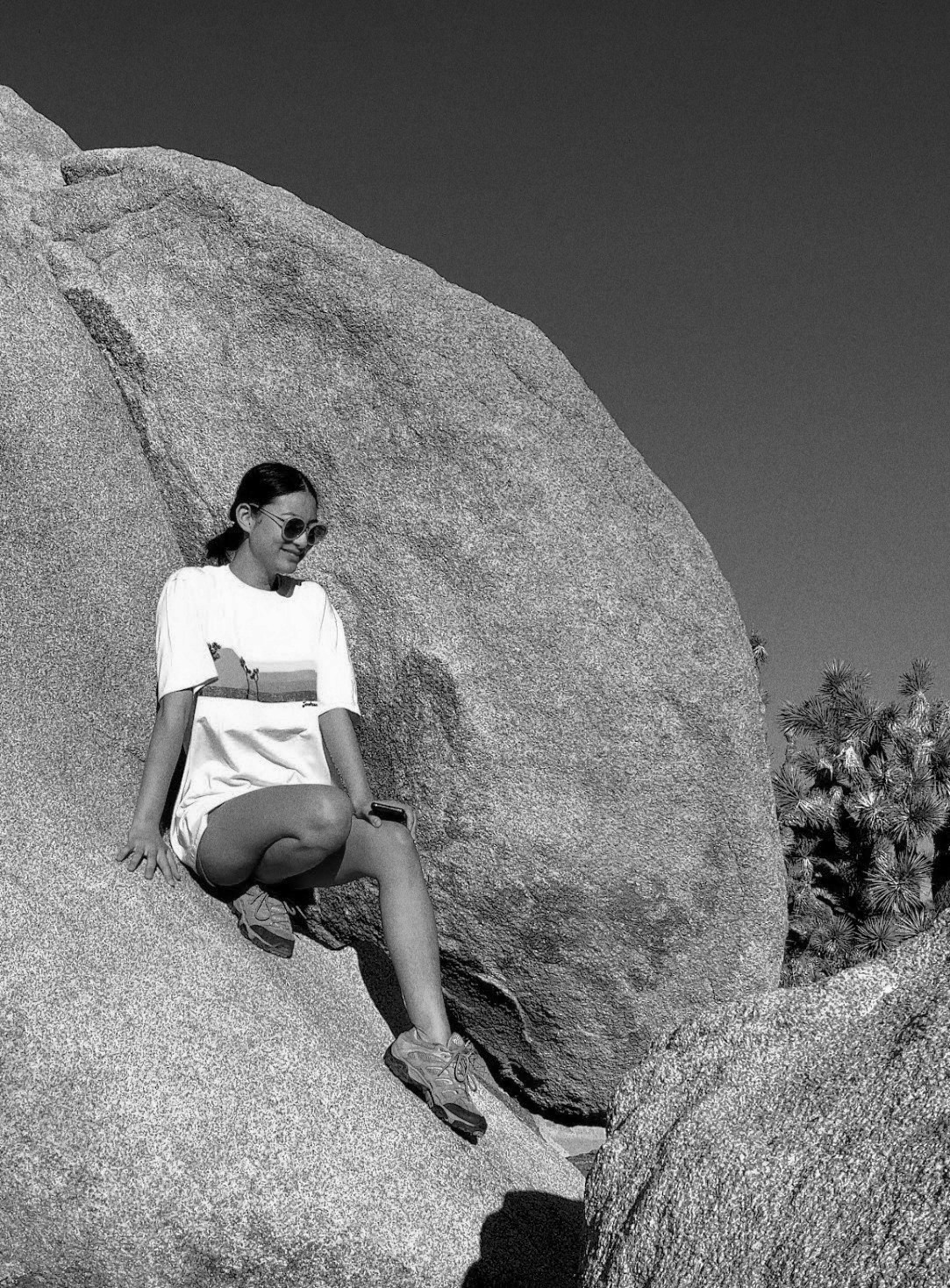 a black-and-white photo of me (alice) sitting on a boulder in the desert, wearing sunglasses and hiking boots.