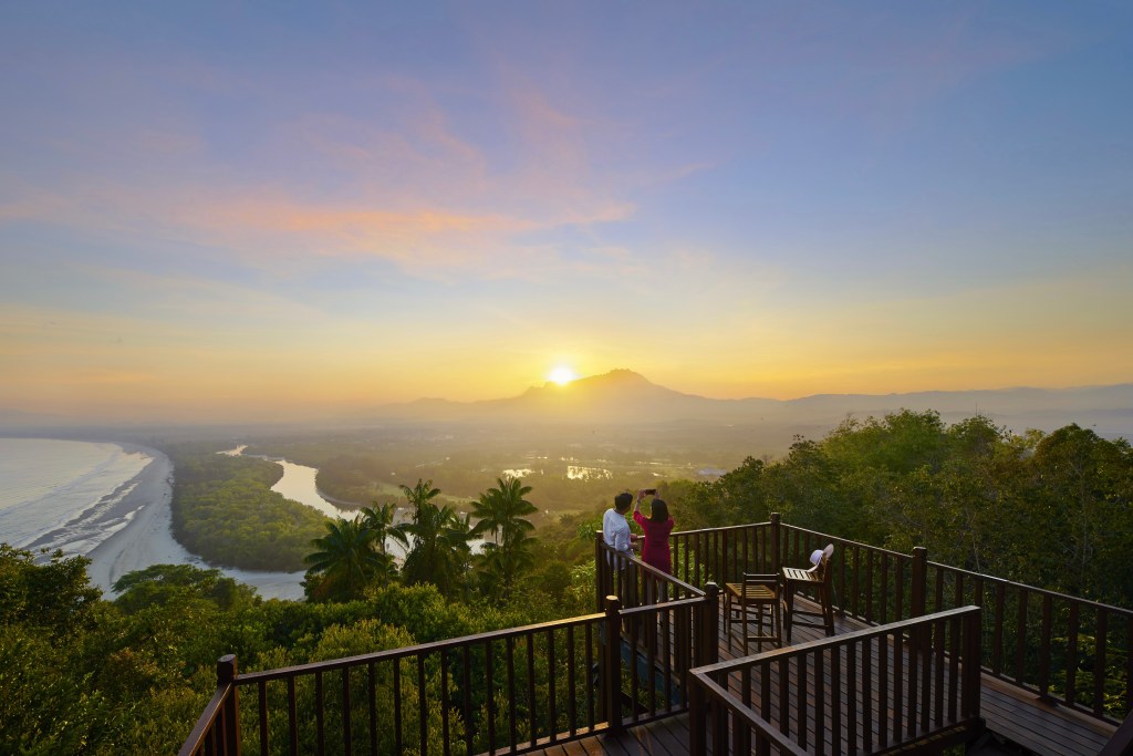 A scenic view of a coastline at sunset, with silhouettes of a couple taking photos from a wooden deck surrounded by lush greenery.