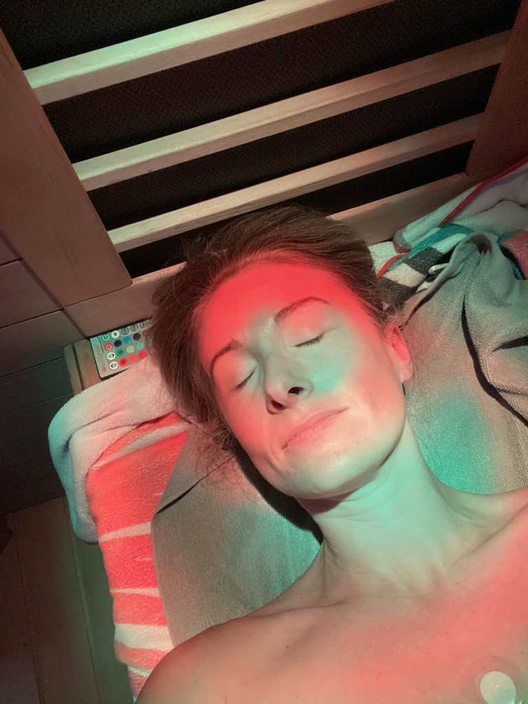 Heather Hausman relaxing under infrared light therapy, lying on a cushion with striped towels and a remote control nearby, bathed in soothing red and turquoise hues.
