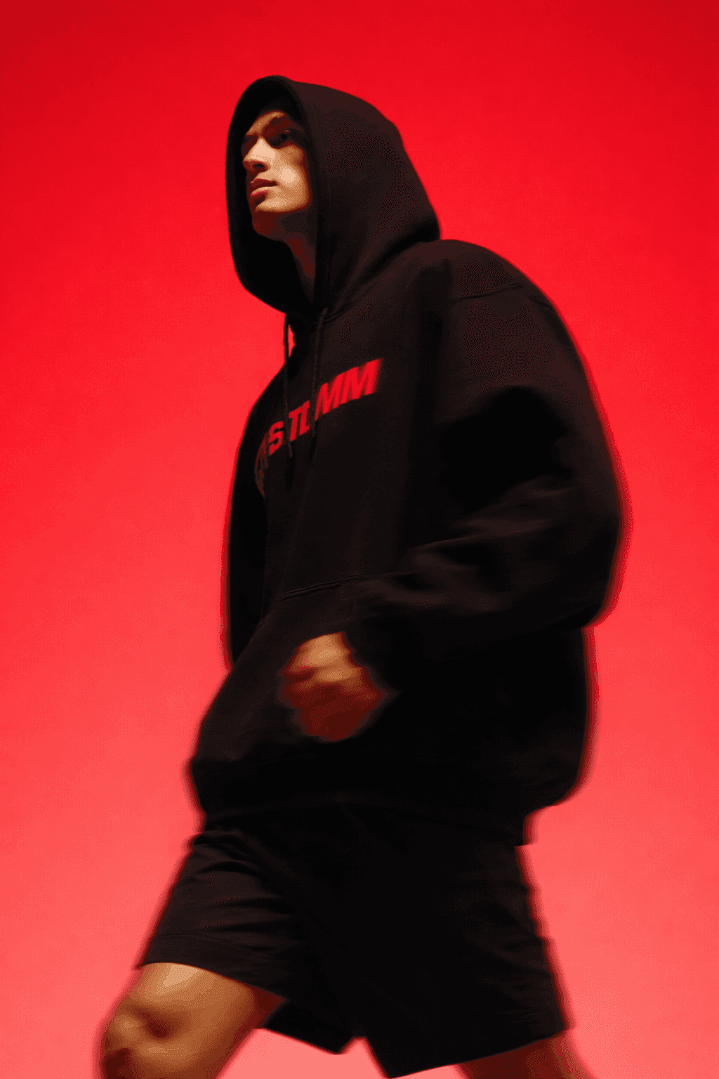 Male model wearing black hoodie with red text walking in motion blur against vibrant red studio background