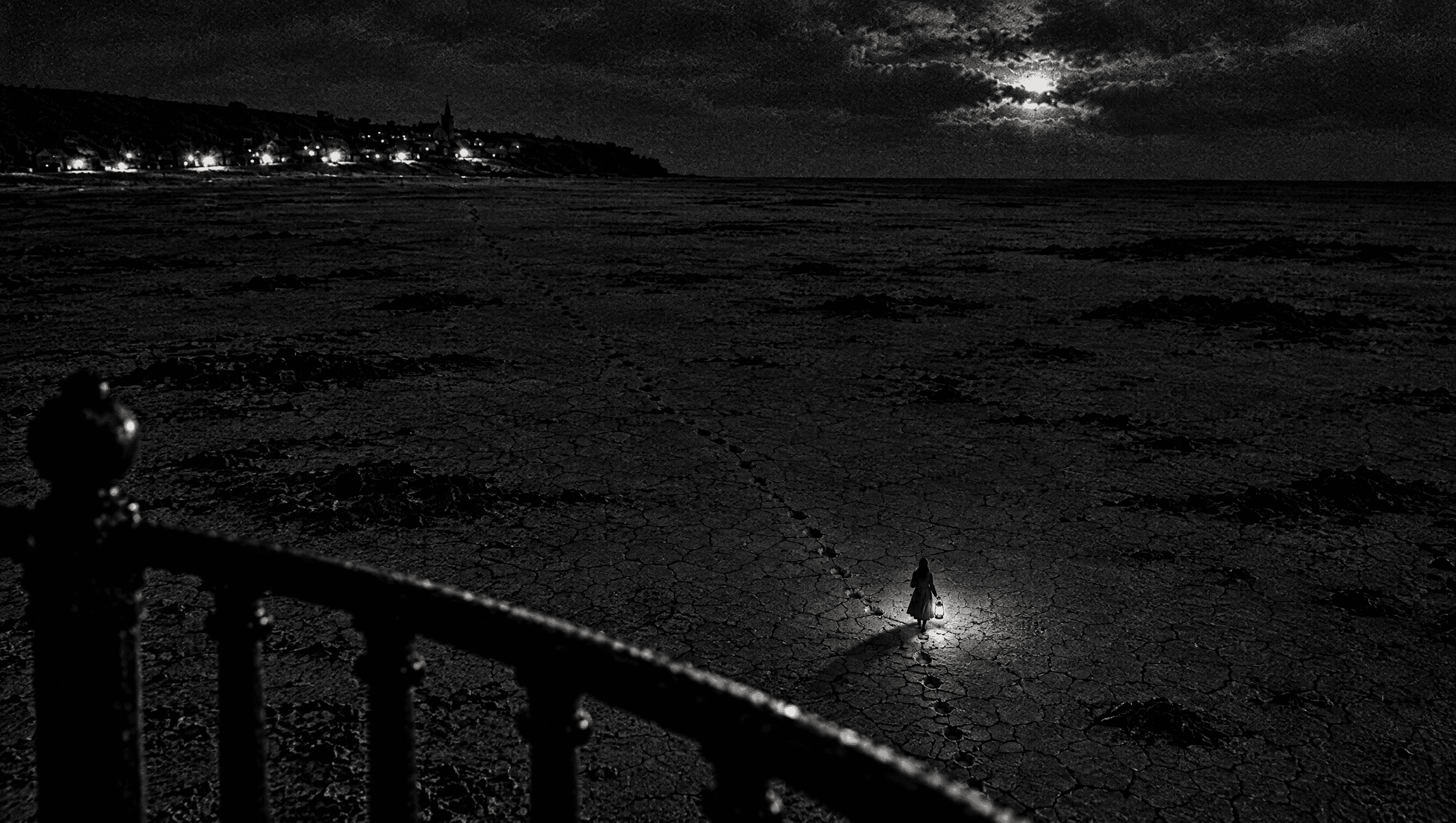 At night, seen from the lighthouse gallery, a woman carries a lantern far out across the dry seabed