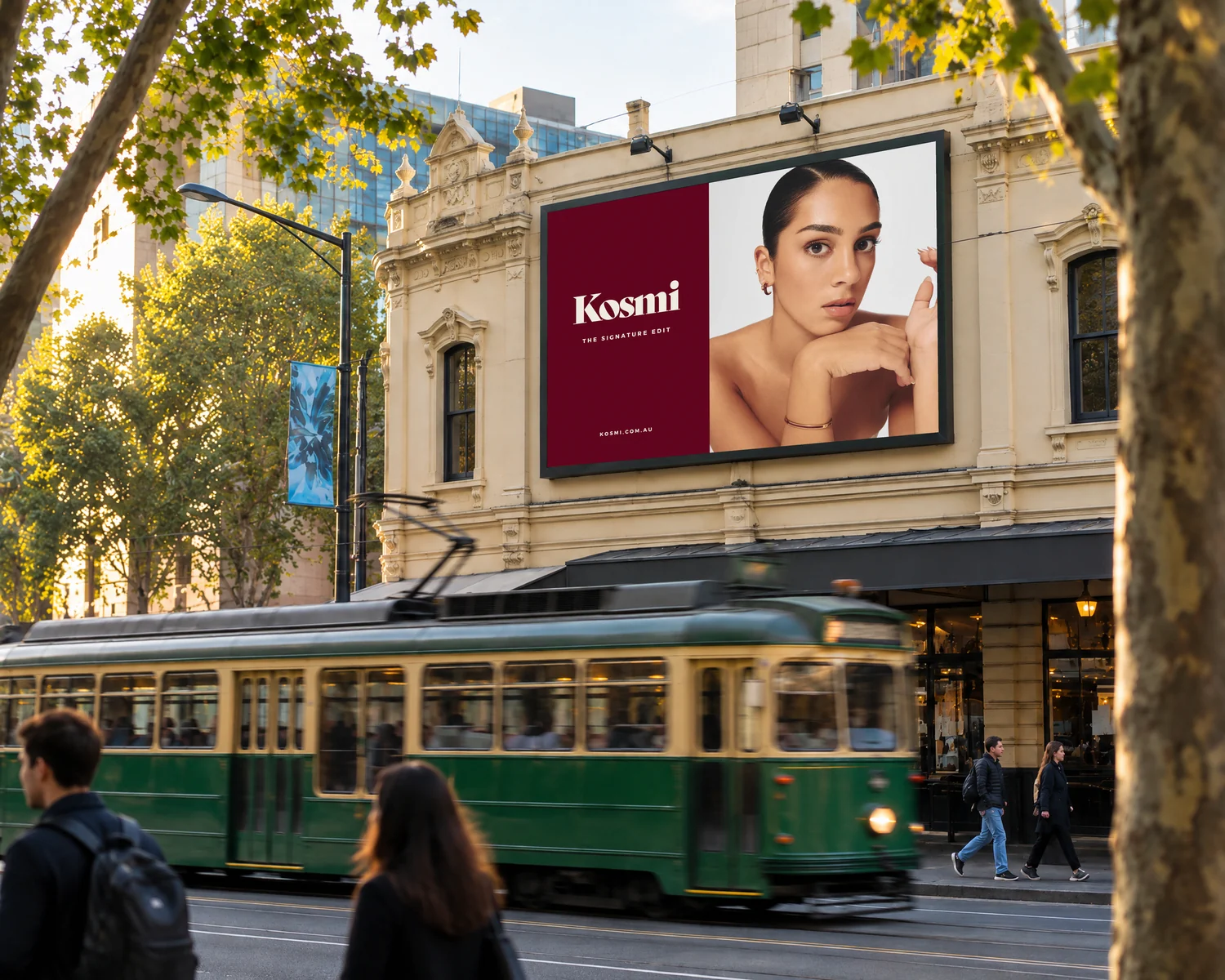 A Kosmi Signature Edit billboard above a city street as a tram passes: a model in gold earrings and a cherry bangle beside the cherry Kosmi logo.