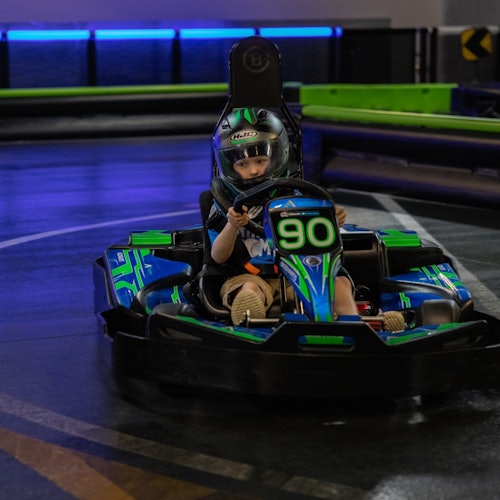 Дете с каска управлява картинг автомобил с номер 90 по осветена с неон вътрешна писта.