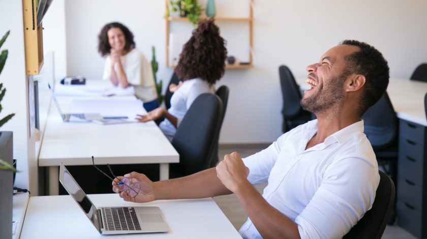 A man is laughing out loud in a office behind his laptop
