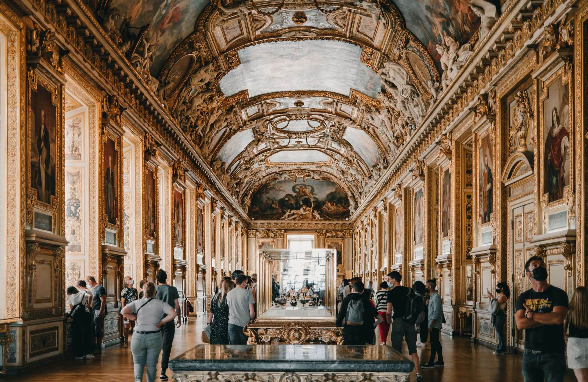 Ornate museum hall with a vaulted, richly decorated ceiling and golden framed paintings. Visitors admire the art, creating a sense of awe and reverence.
