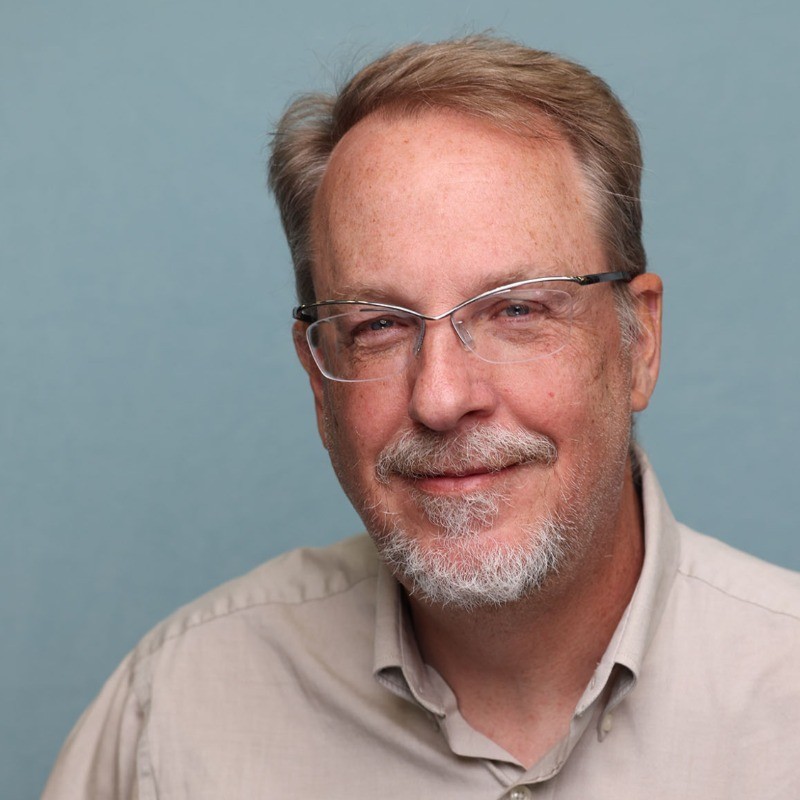 A portrait of a white man with greying blond hair and silver glasses, wearing a tan collared shirt