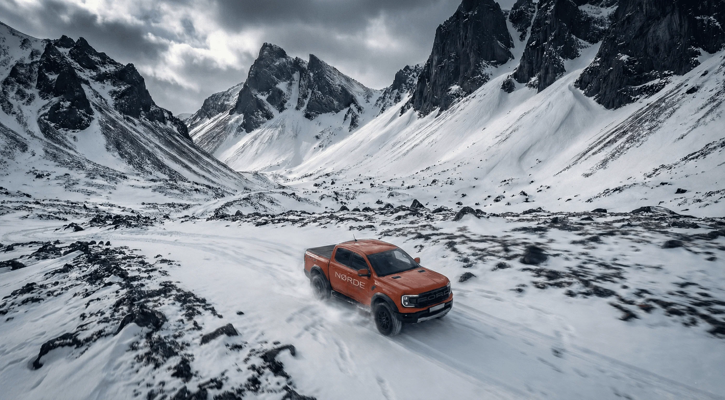 An orange Ford Ranger Raptor driving along a snowy track through a valley surrounded by white mountains.