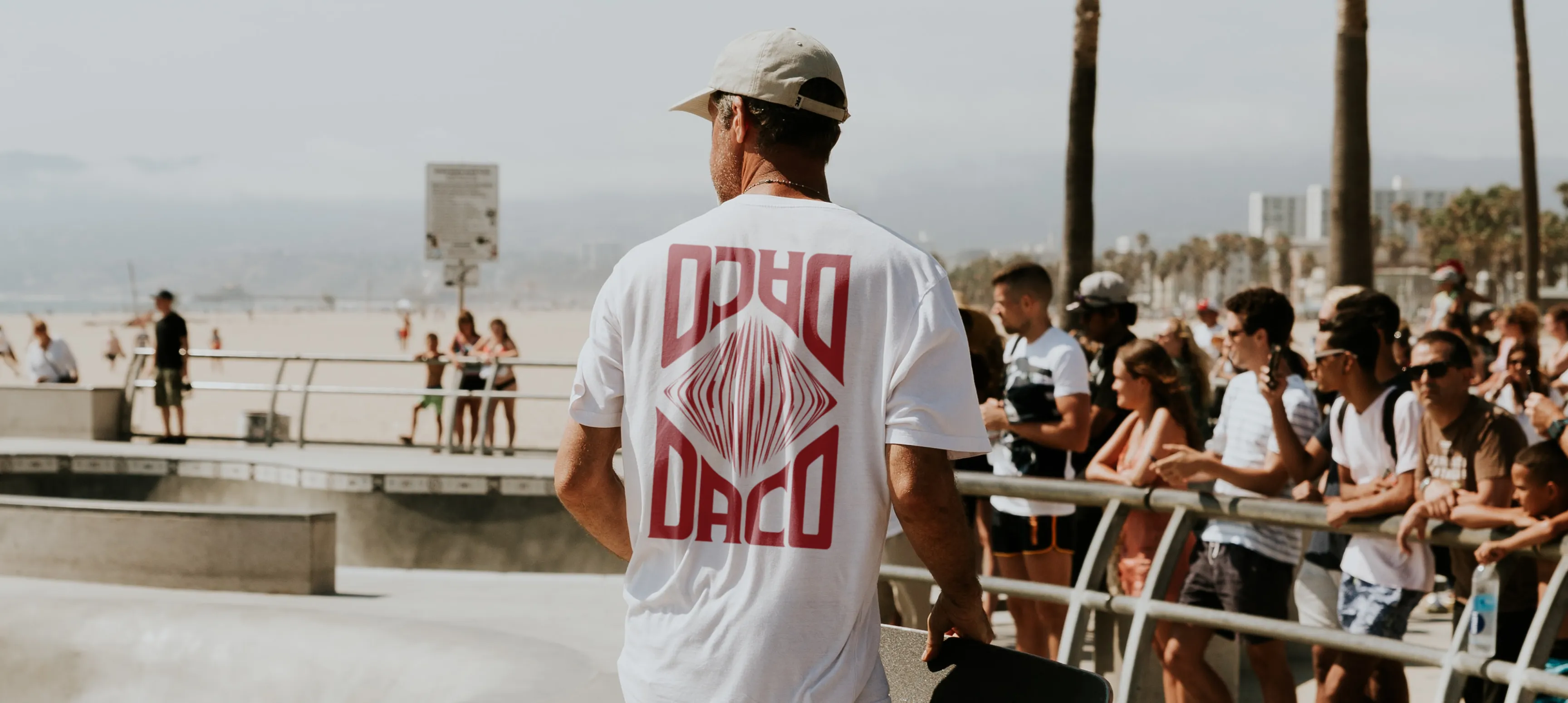 A man skateboarding at a skatepark wearing a DACO white tshirt. The tshirt has the DACO wordmark reflected top and bottom with a diamond in the middle. 