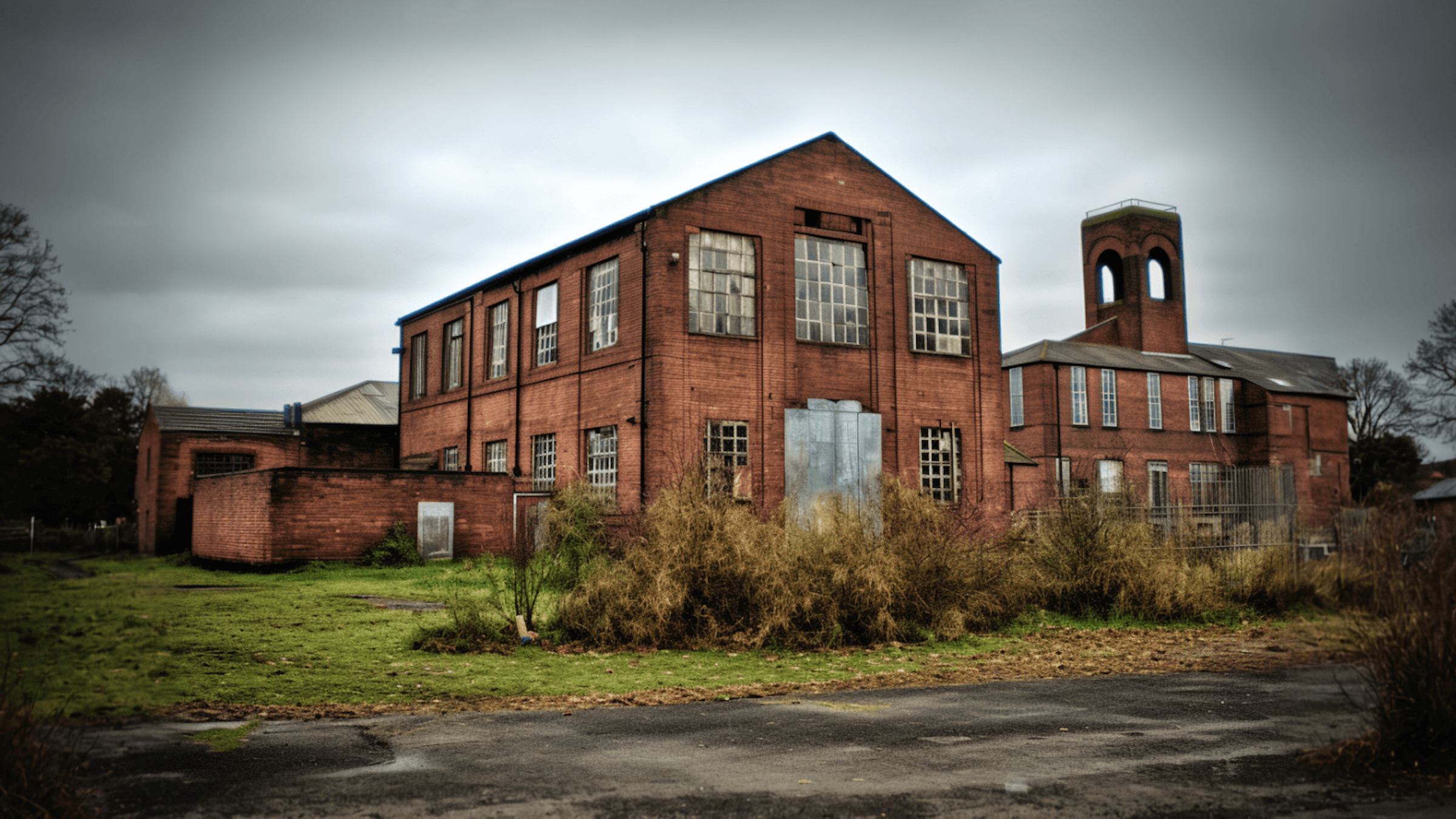 Abandoned red brick factory building surrounded by overgrown grass under a cloudy sky.