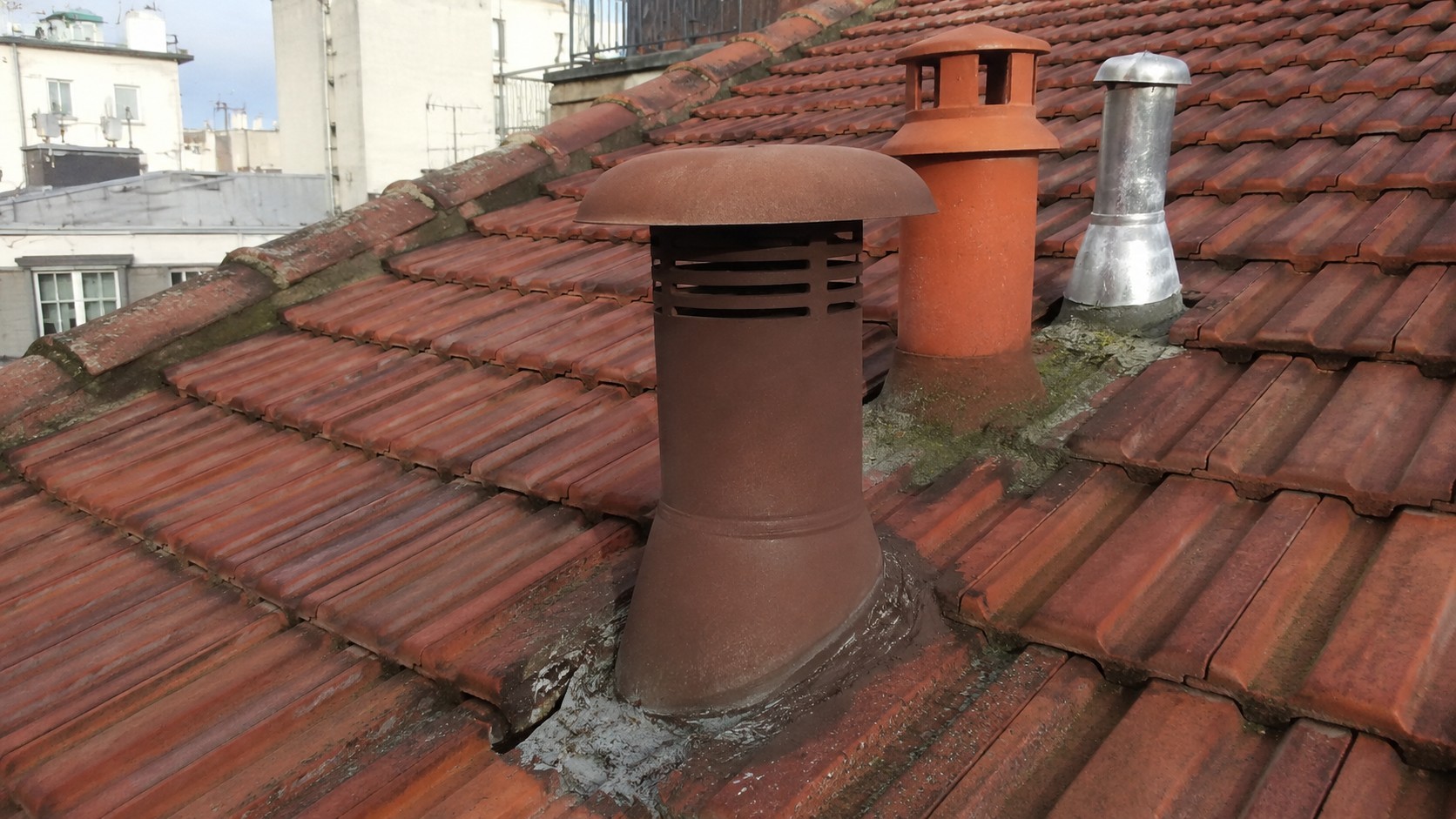Vue rapprochée d’une toiture parisienne en tuiles terre cuite équipée de plusieurs sorties techniques de toiture, dont une tuile à douille de ventilation, un conduit métallique d’extraction et une sortie d’aération, assurant la ventilation et l’évacuation des réseaux techniques du bâtiment.