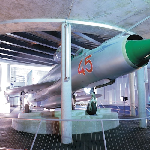 Ett silverfärgat stridsflygplan med en grön noskon och nummer 45 visat, utställt i ett inomhusmuseum bakom räcken.