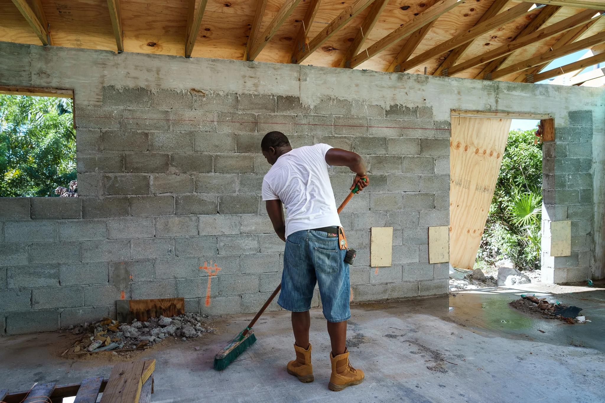 A person wearing a white shirt, denim shorts, and boots is sweeping debris on a concrete floor inside a partially constructed building with wooden beams and cinder block walls. Theres an opening to the outside with trees visible.