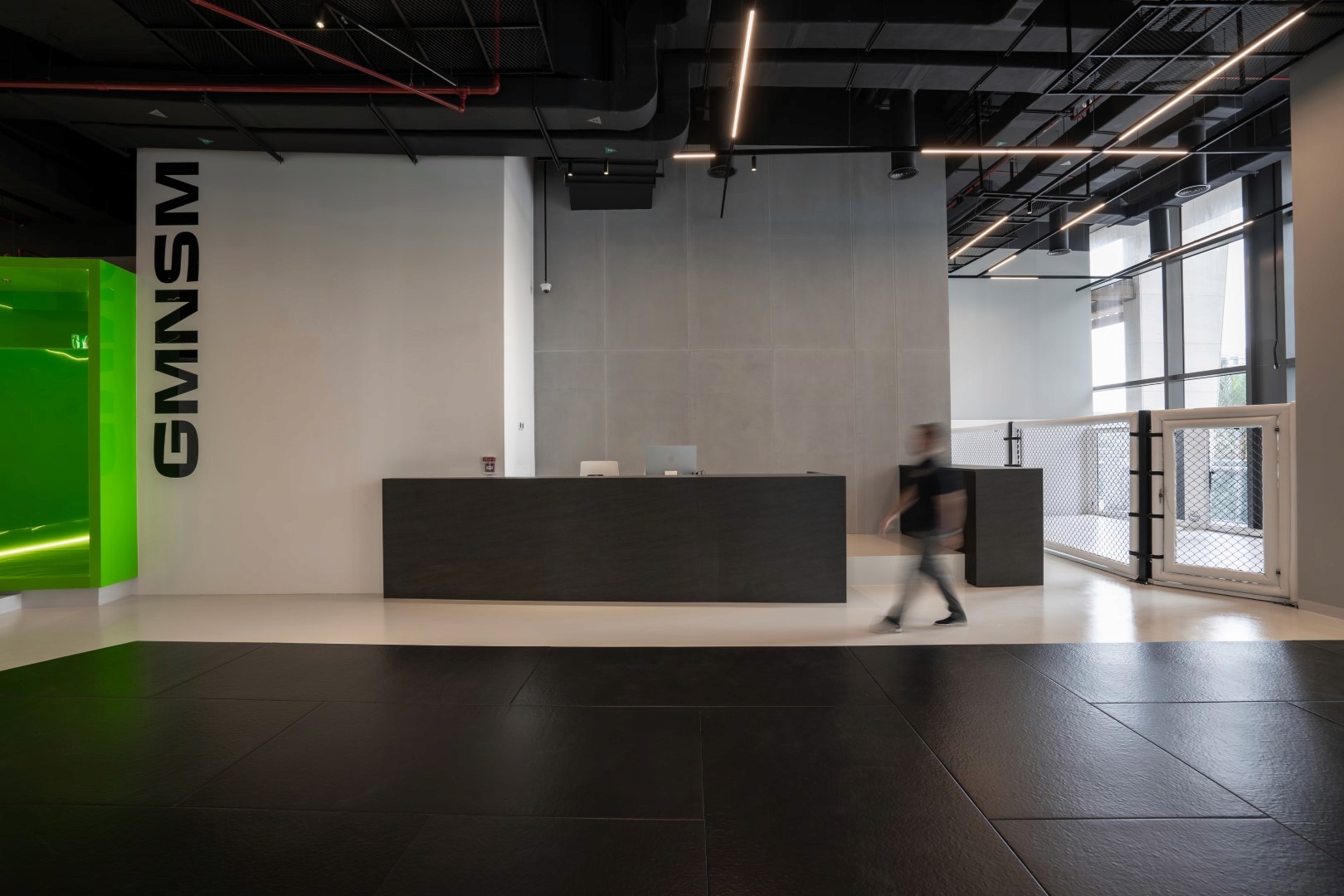 A modern gym reception area features a sleek black and white design with a minimalist counter, bright green accent wall, and overhead industrial-style lighting.