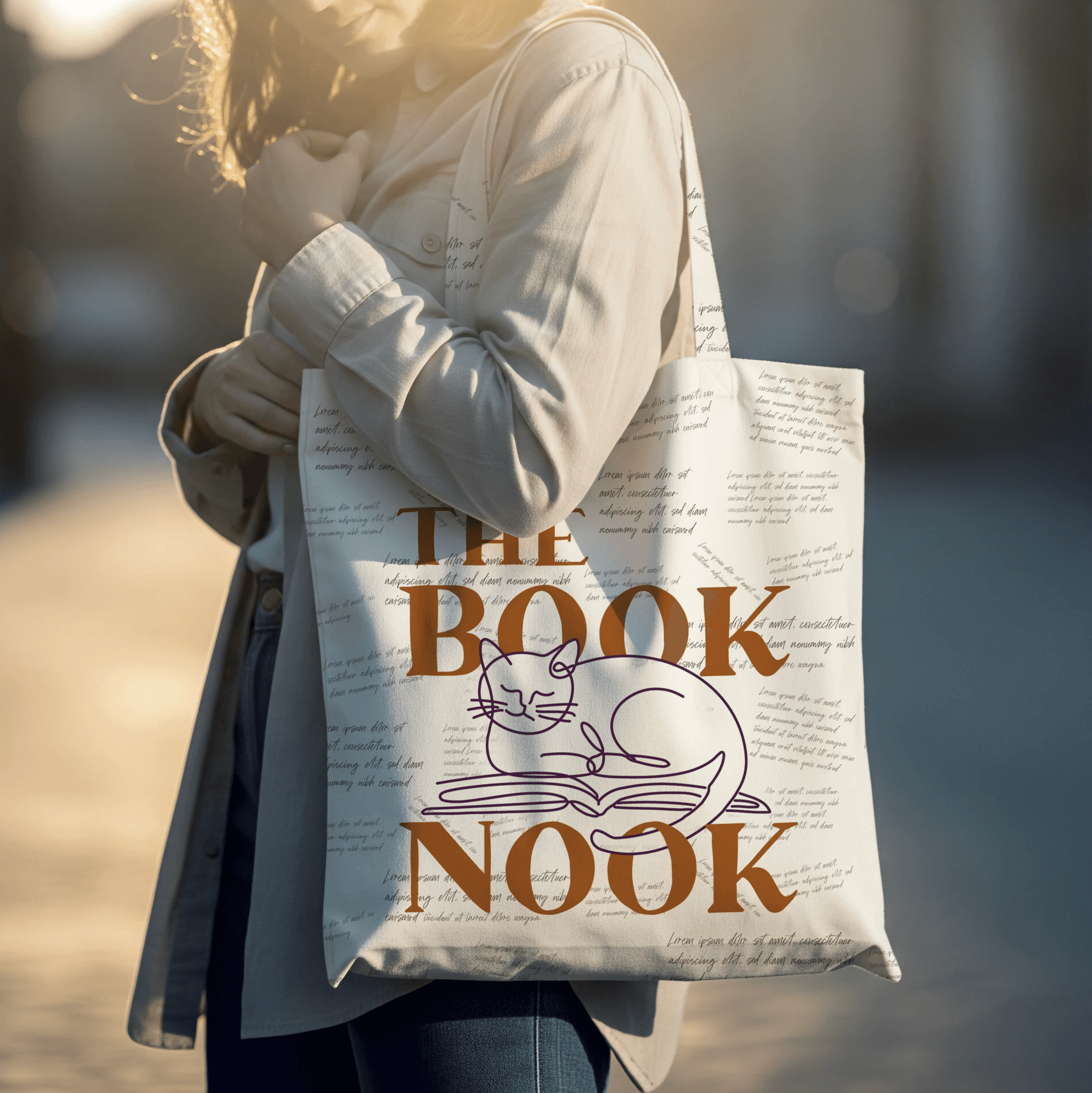 A canvas tote bag printed with THE BOOKNOOK in serif letters and a line-art cat, with a pattern of quotes in a handwritten font, carried outdoors by a woman in a cardigan.