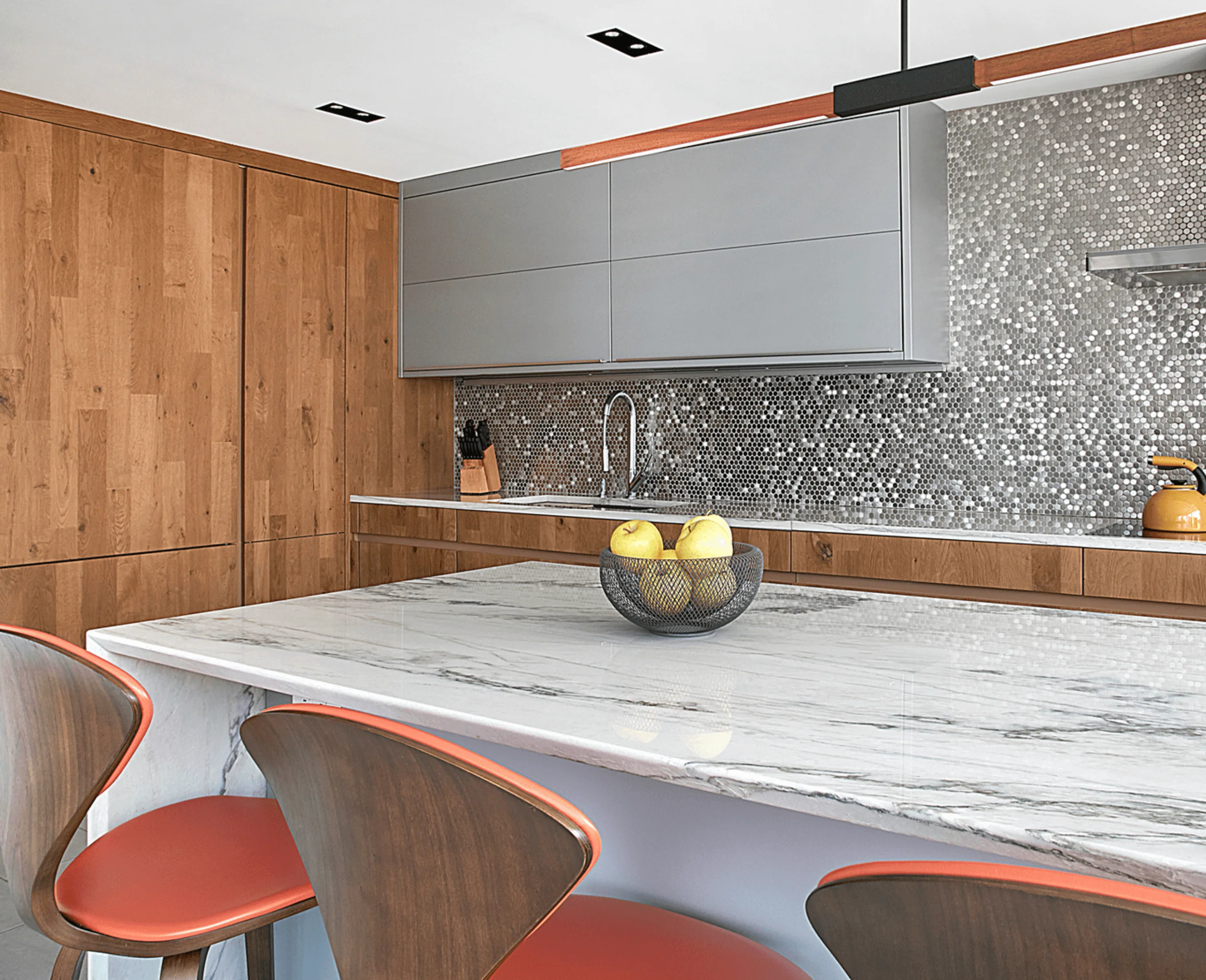 A close-up of the kitchen's white marble island with a bowl of apples, walnut cherner stools, and a mirrored penny-tile backsplash