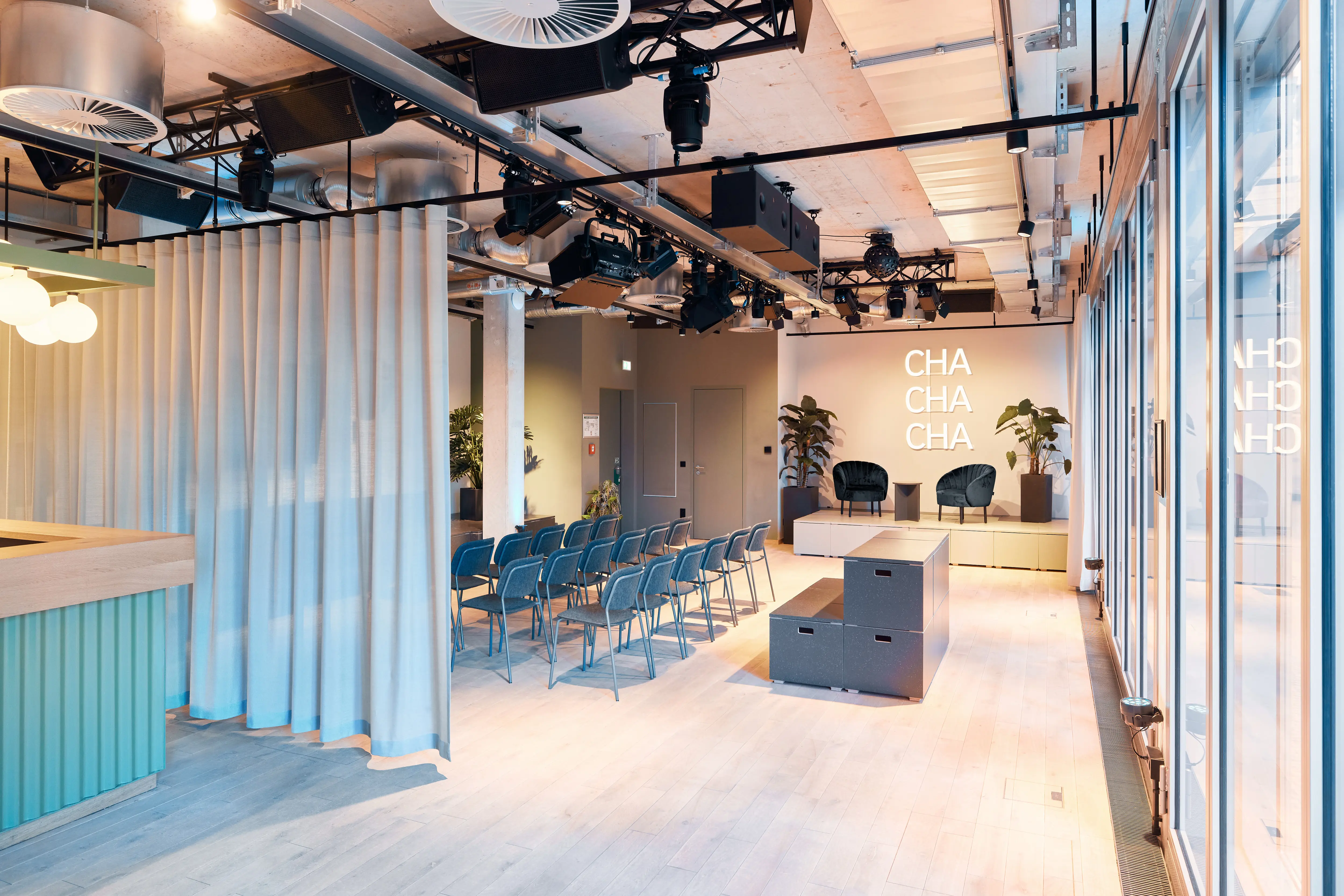 Event room at 381 EVENT SPACE divided by a light blue curtain, with rows of blue chairs, grey modular seating cubes and the illuminated CHA CHA CHA wall.