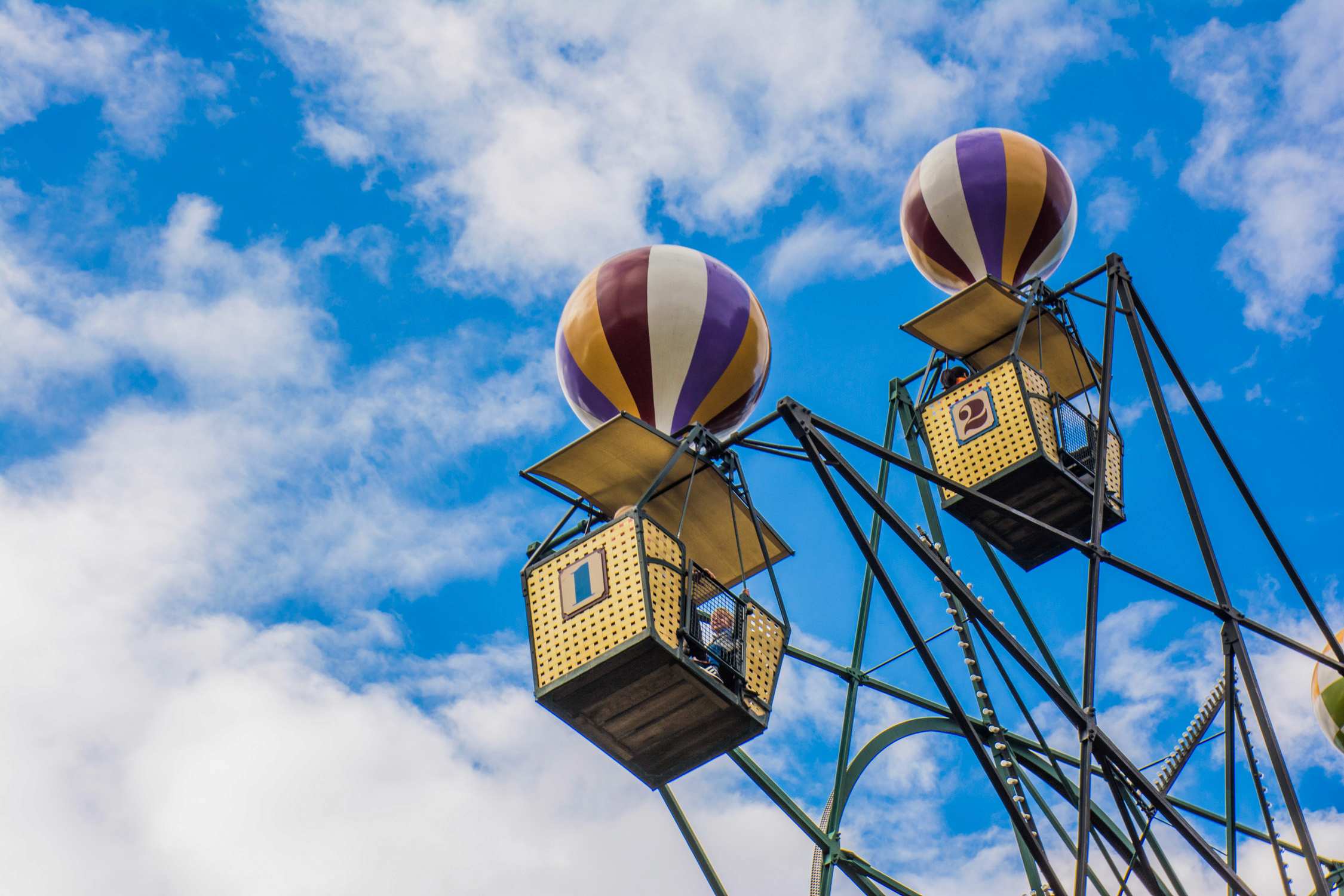 Two hot air balloon-shaped gondolas on a tall amusement park ride against a blue sky with white clouds.