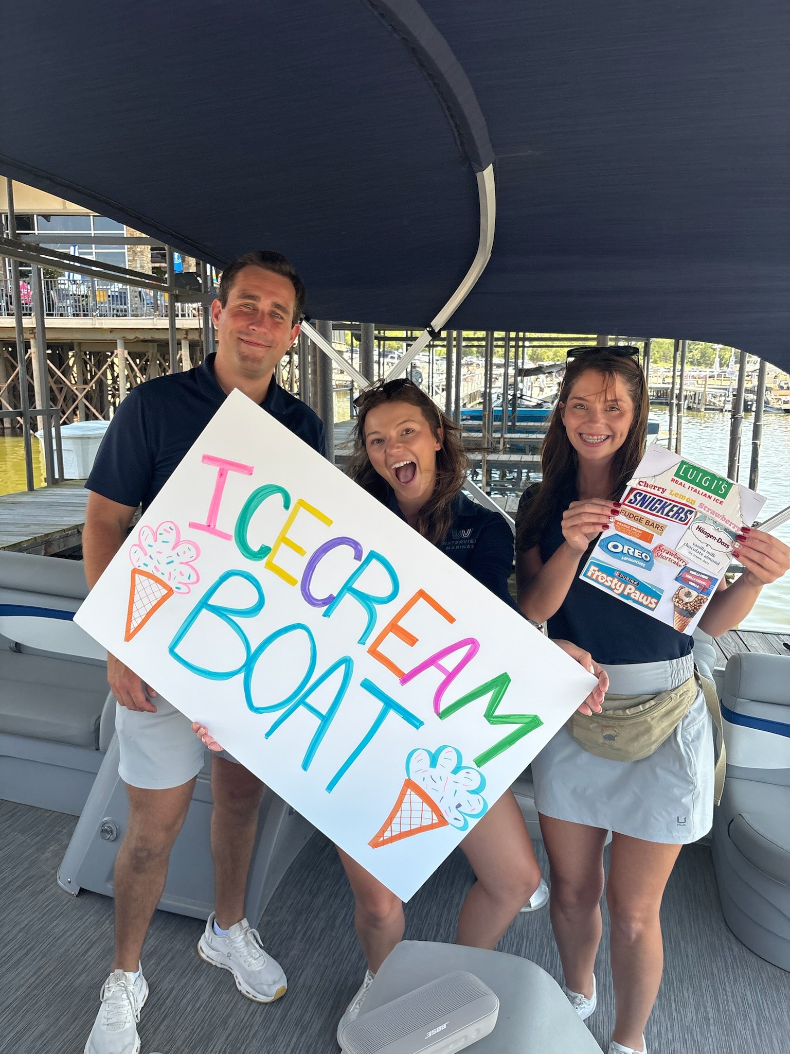 Three people on a boat hold a colorful sign that reads "ICE CREAM BOAT" along with ice cream cone illustrations, with one person also displaying various ice cream brand labels.