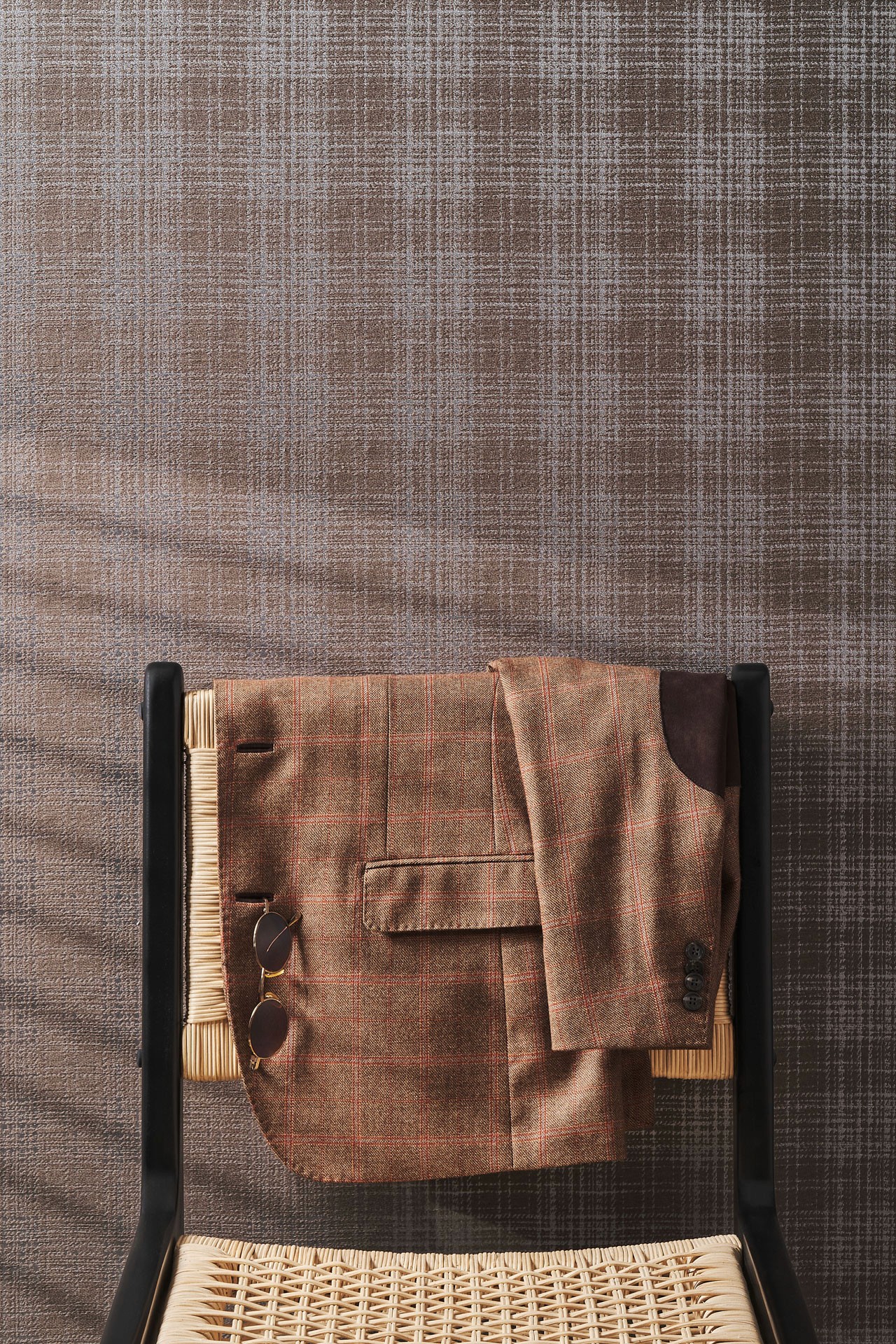 Giacca sartoriale marrone a quadri appoggiata sullo schienale di una sedia in legno con seduta intrecciata; un paio di occhiali da sole è agganciato alla tasca, sullo sfondo di una parete tessile nei toni neutri.