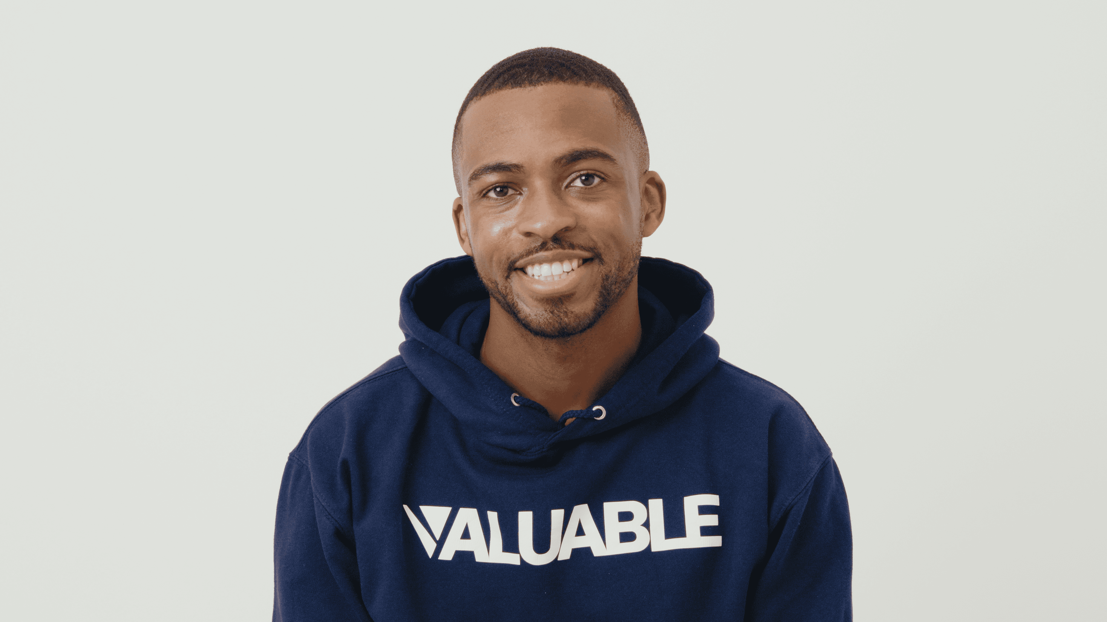 Man in a navy blue valuable hoodie with the word VALUABLE printed on it smiles.